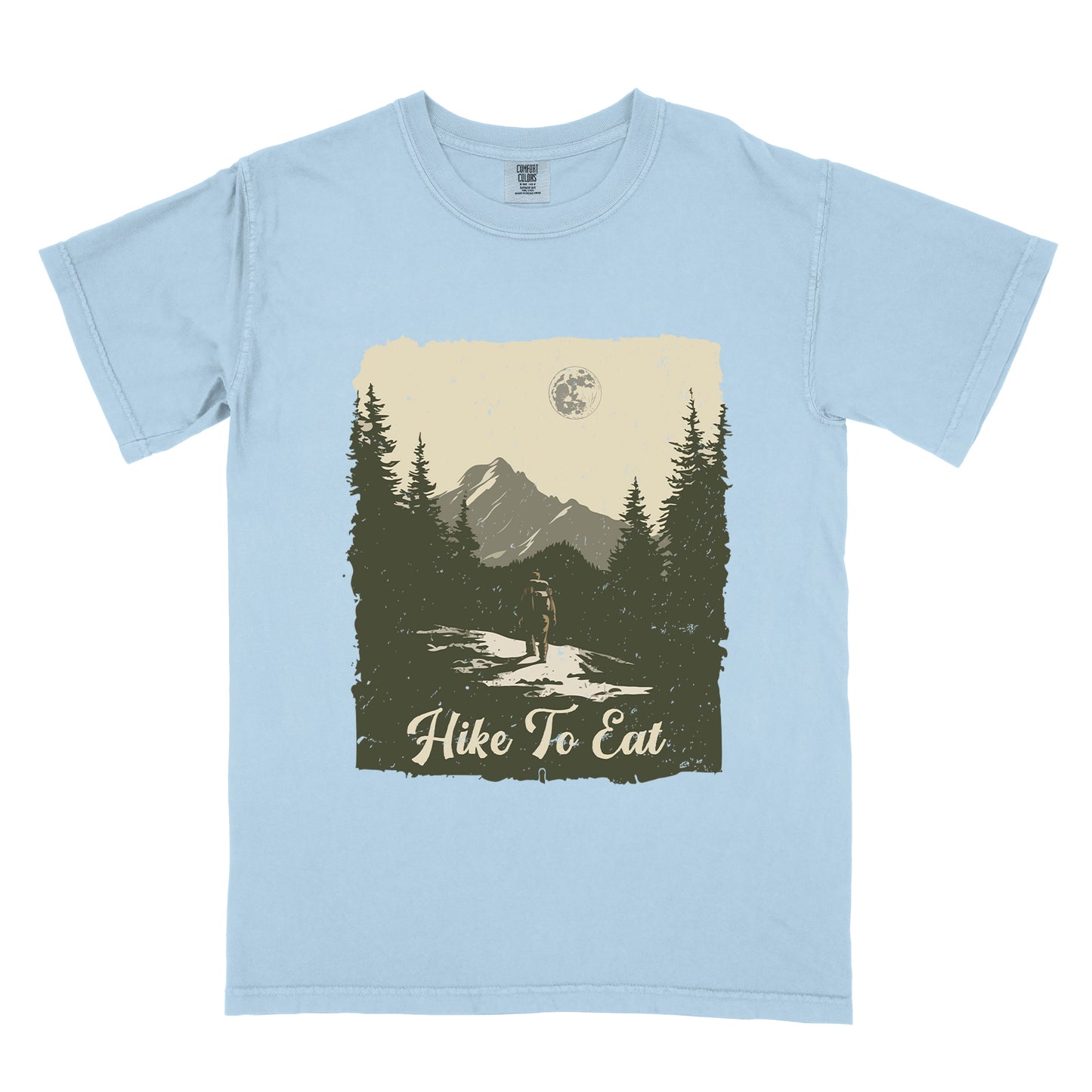 Chambray (Light Blue) t-shirt featuring a vintage-style illustration of a lone hiker on a mountain trail. The rectangular design shows pine trees, mountain silhouettes, and a full moon against a cream-colored sky. A solitary hiker with backpack stands on the path facing the mountains. "Hike To Eat" appears in cream script at the bottom of the illustration. The artwork uses muted green, cream, and gray tones in a distressed, rustic style.