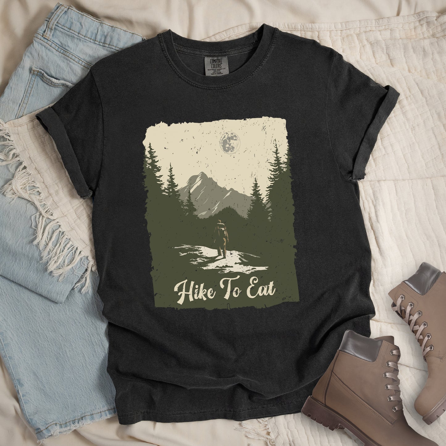 Graphite (Deep Gray) t-shirt featuring a vintage-style illustration of a lone hiker on a mountain trail. The rectangular design shows pine trees, mountain silhouettes, and a full moon against a cream-colored sky. A solitary hiker with backpack stands on the path facing the mountains. "Hike To Eat" appears in cream script at the bottom of the illustration. The artwork uses muted green, cream, and gray tones in a distressed, rustic style.