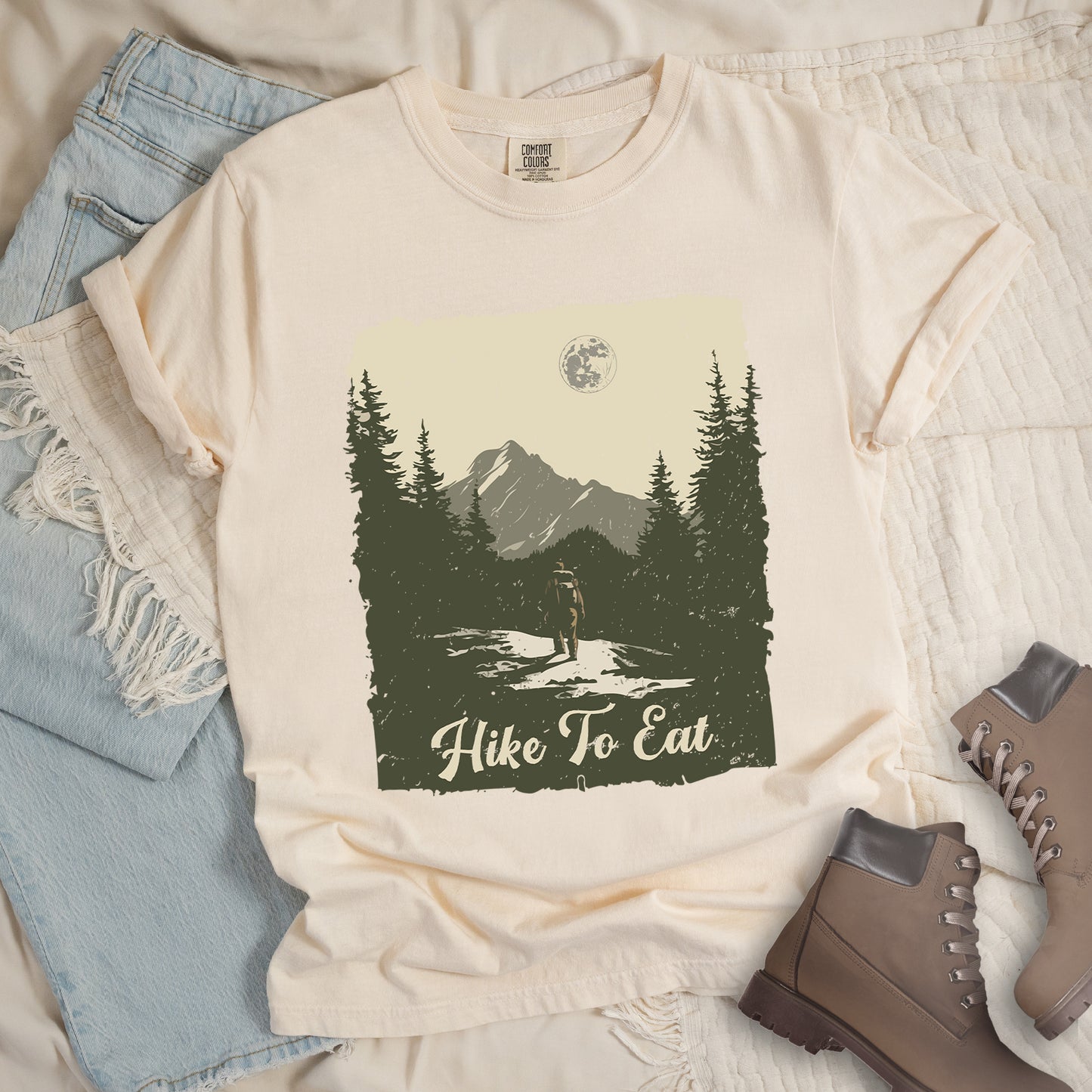 Ivory (Cream White) t-shirt featuring a vintage-style illustration of a lone hiker on a mountain trail. The rectangular design shows pine trees, mountain silhouettes, and a full moon against a cream-colored sky. A solitary hiker with backpack stands on the path facing the mountains. "Hike To Eat" appears in cream script at the bottom of the illustration. The artwork uses muted green, cream, and gray tones in a distressed, rustic style.