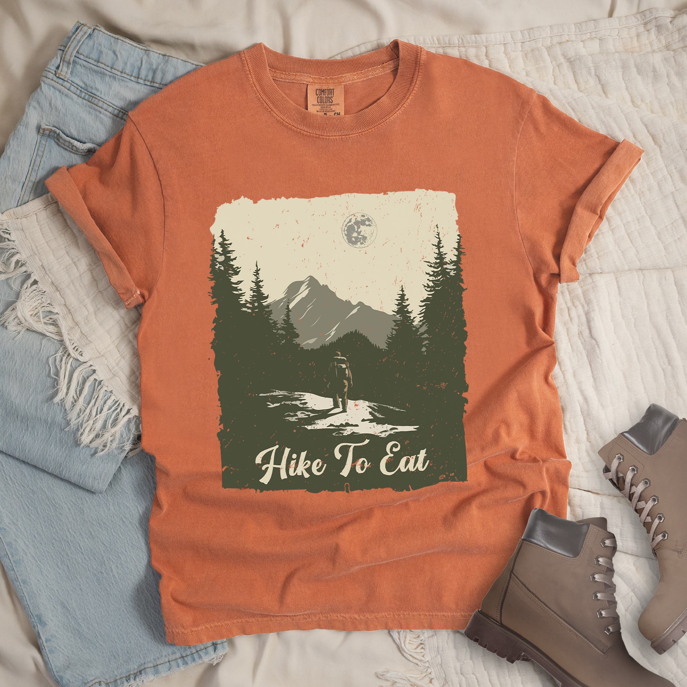 Orange shirt with Hiking Design featuring the phrase “Hike to Eat” and an illustration of a lone hiker walking toward mountains under the moonlight in a nature scene.