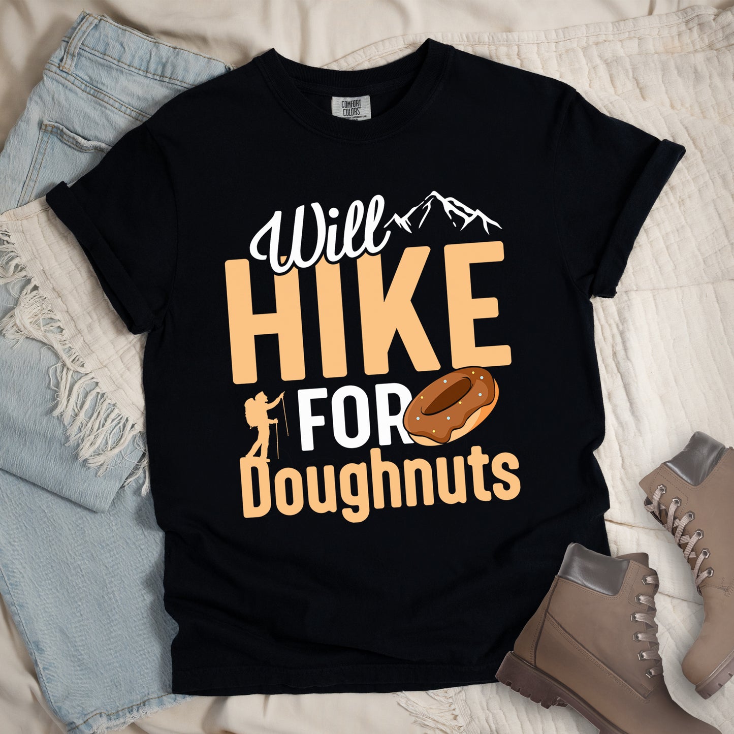 Black t-shirt with "Will HIKE FOR Doughnuts" graphic design. White script "Will" above tan "HIKE" in bold letters, with small mountain peaks above. "FOR" appears in white, followed by "Doughnuts" in tan, with an illustrated chocolate donut replacing the "O" in "FOR". Features a small hiker silhouette. The shirt features a classic crew neck and casual rolled sleeves, styled alongside light wash denim and brown hiking boots.