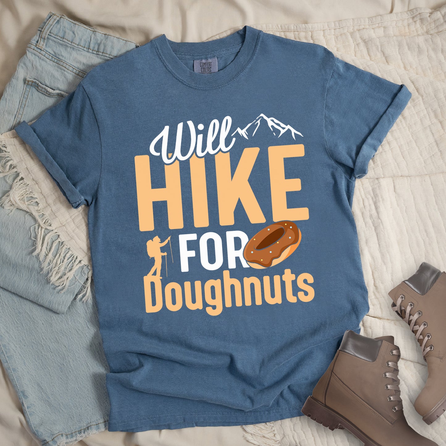Blue Jean(Blue) t-shirt with "Will HIKE FOR Doughnuts" graphic design. White script "Will" above tan "HIKE" in bold letters, with small mountain peaks above. "FOR" appears in white, followed by "Doughnuts" in tan, with an illustrated chocolate donut replacing the "O" in "FOR". Features a small hiker silhouette. The shirt features a classic crew neck and casual rolled sleeves, styled alongside light wash denim and brown hiking boots. Perfect for hiking enthusiasts with a sweet tooth who enjoy post-trek sweet