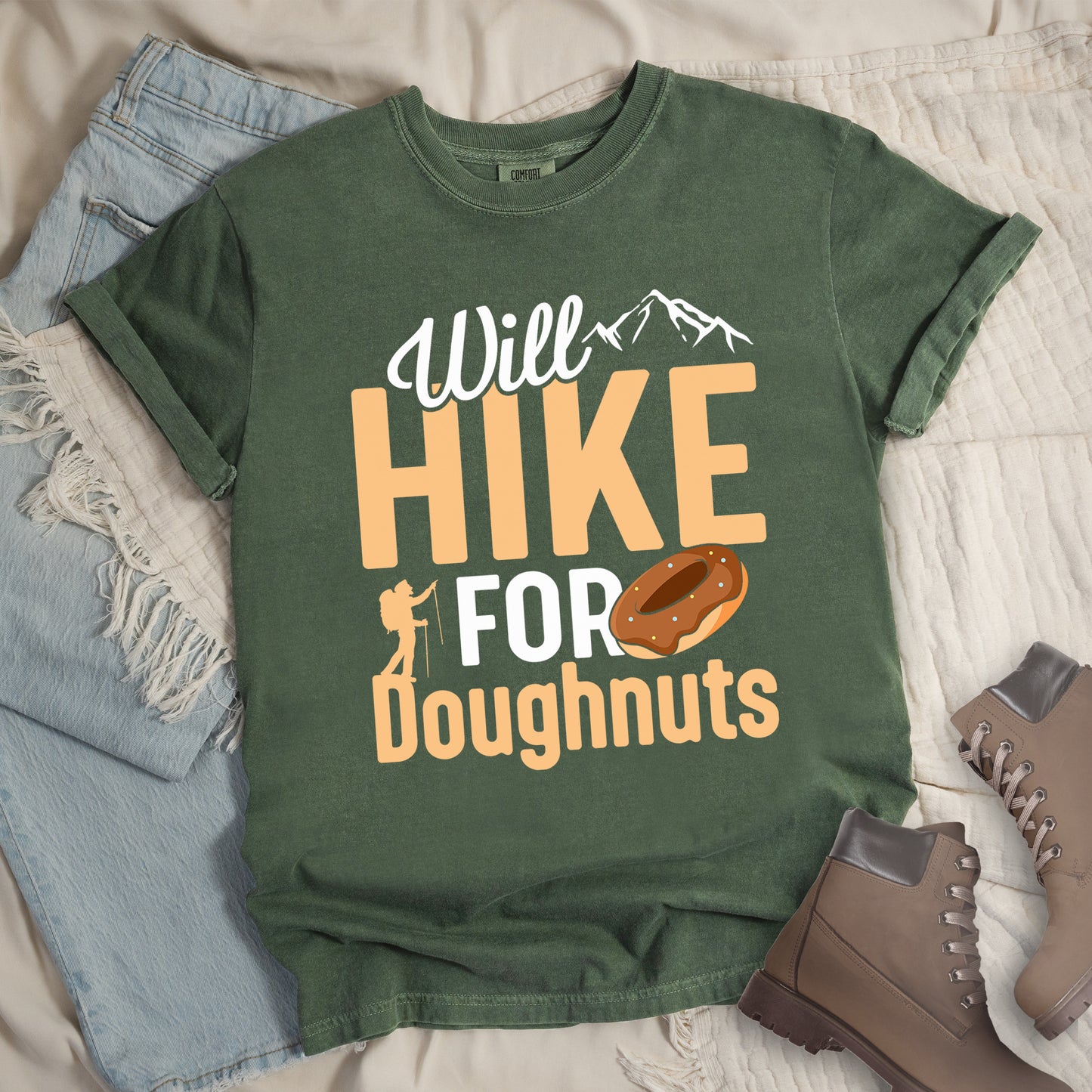 Moss (Green) t-shirt with "Will HIKE FOR Doughnuts" graphic design. White script "Will" above tan "HIKE" in bold letters, with small mountain peaks above. "FOR" appears in white, followed by "Doughnuts" in tan, with an illustrated chocolate donut replacing the "O" in "FOR". Features a small hiker silhouette. The shirt features a classic crew neck and casual rolled sleeves, styled alongside light wash denim and brown hiking boots.