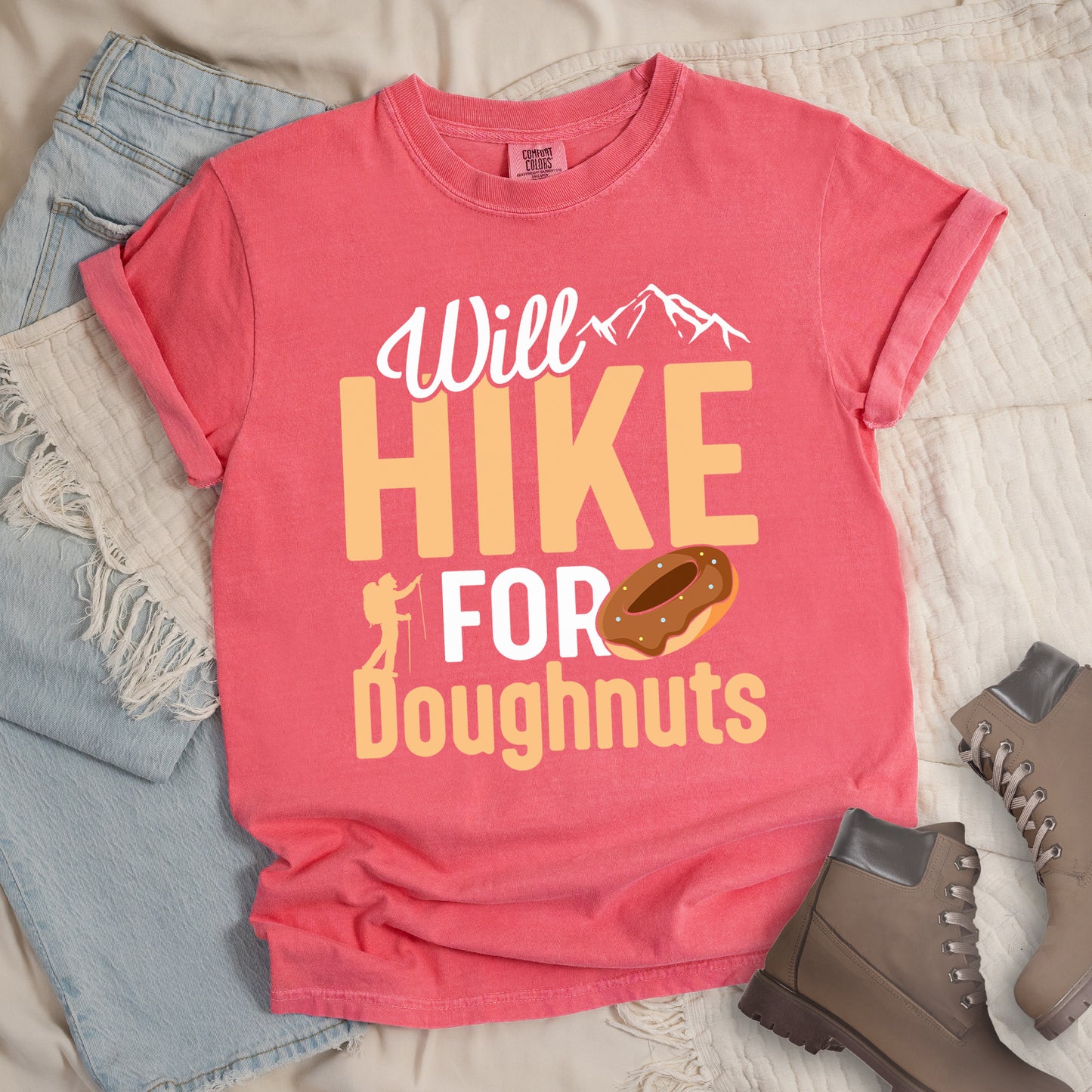 Watermelon (Light Coral) t-shirt with "Will HIKE FOR Doughnuts" graphic design. White script "Will" above tan "HIKE" in bold letters, with small mountain peaks above. "FOR" appears in white, followed by "Doughnuts" in tan, with an illustrated chocolate donut replacing the "O" in "FOR". Features a small hiker silhouette. The shirt features a classic crew neck and casual rolled sleeves, styled alongside light wash denim and brown hiking boots.