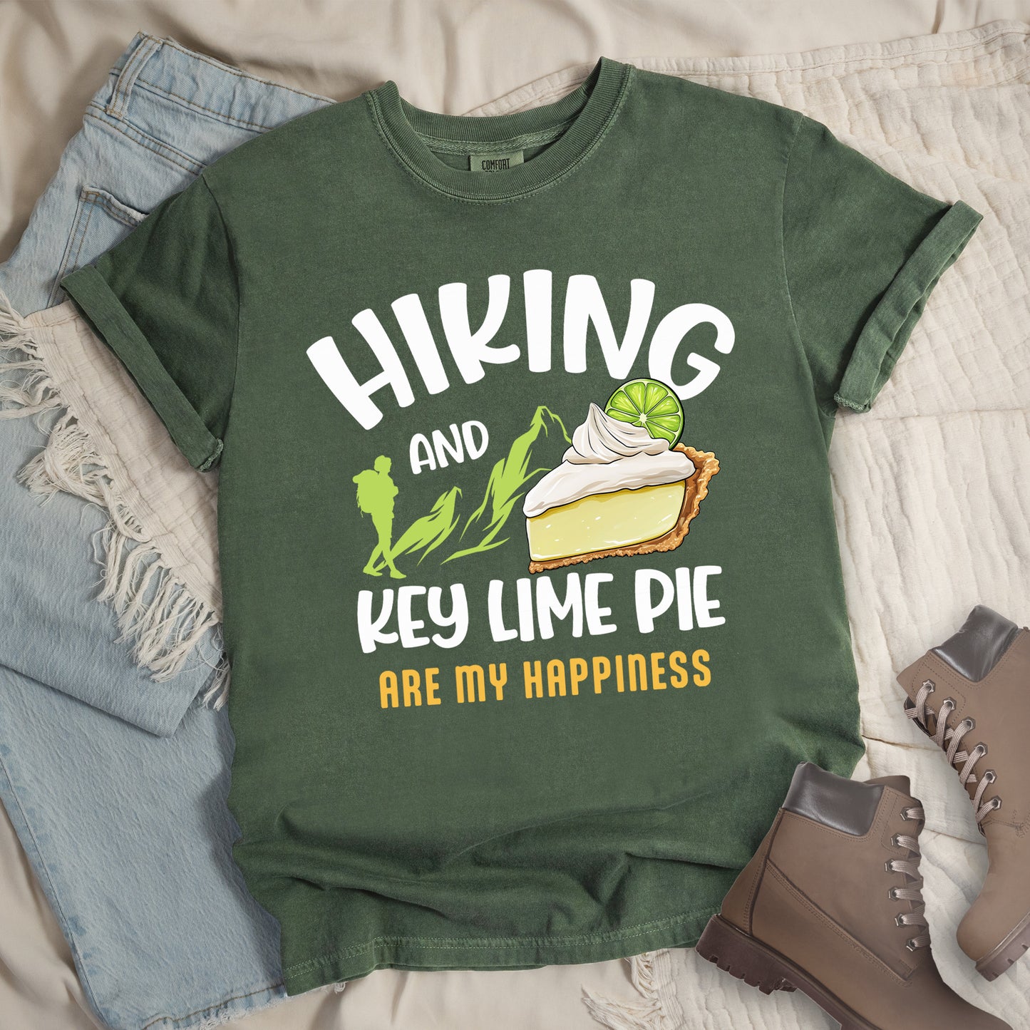 Moss (Dark Green) t-shirt with text "HIKING AND KEY LIME PIE ARE MY HAPPINESS". "HIKING" appears in white arched letters at the top, "AND" in small white text, "KEY LIME PIE" in large white text in the middle, and "ARE MY HAPPINESS" in gold/yellow at the bottom. The design includes green mountain silhouettes with a hiker silhouette and an illustration of a slice of key lime pie with whipped cream and lime garnish in between "AND" and "KEY LIME PIE."