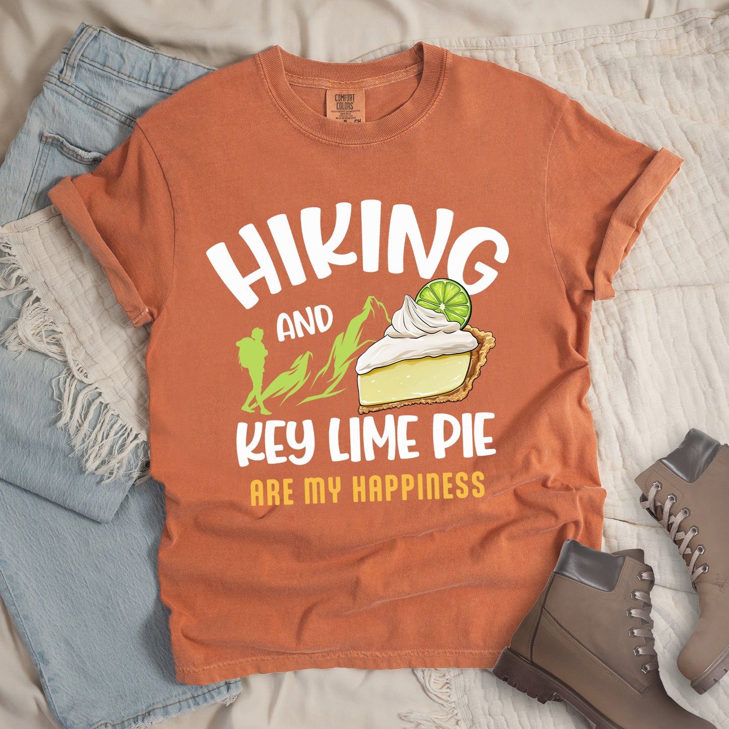Yam (Brown) t-shirt with text "HIKING AND KEY LIME PIE ARE MY HAPPINESS". "HIKING" appears in white arched letters at the top, "AND" in small white text, "KEY LIME PIE" in large white text in the middle, and "ARE MY HAPPINESS" in gold/yellow at the bottom. The design includes green mountain silhouettes with a hiker silhouette and an illustration of a slice of key lime pie with whipped cream and lime garnish in between "AND" and "KEY LIME PIE."