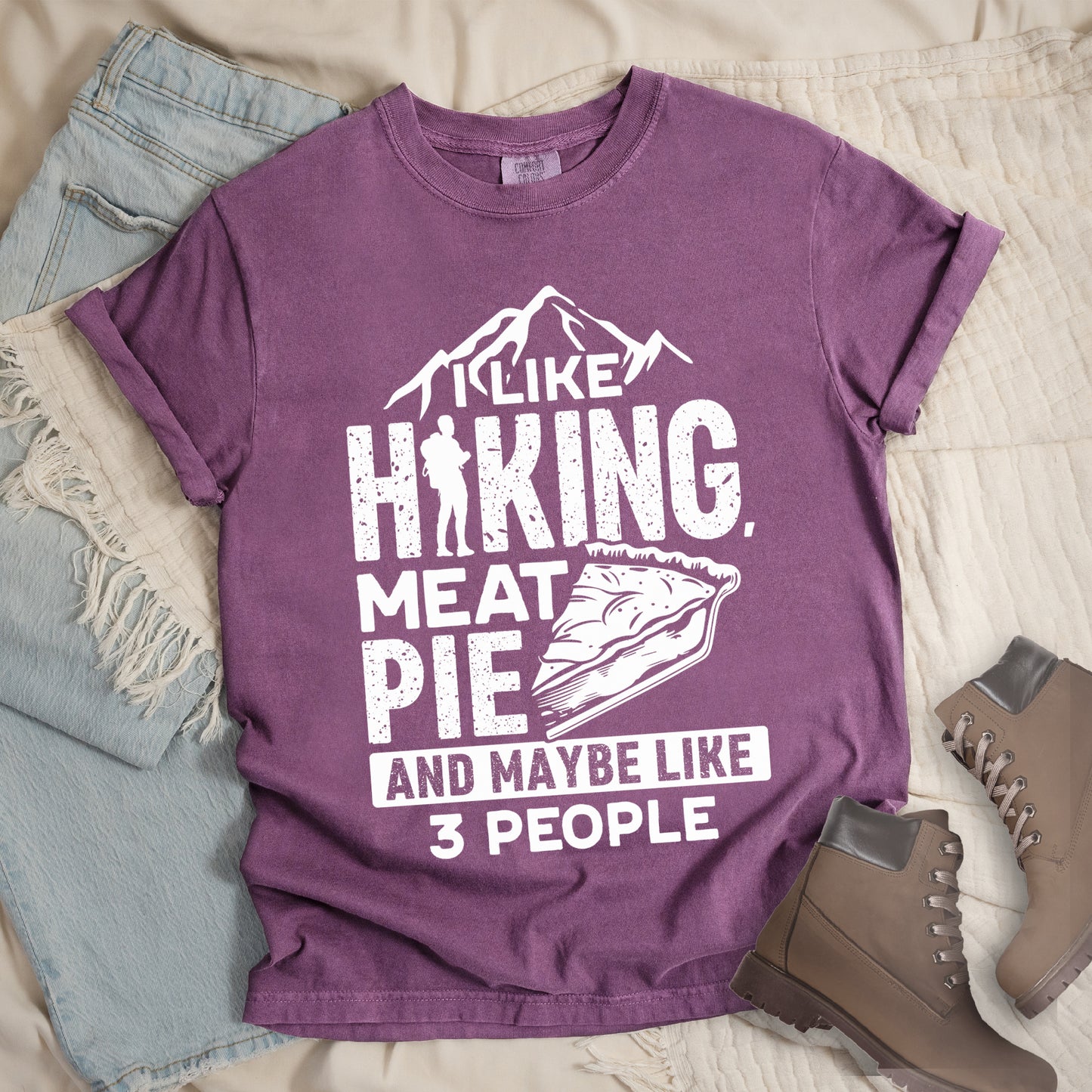Berry (Dark Violet) t-shirt with white distressed text reading "I LIKE HIKING, MEAT PIE AND MAYBE LIKE 3 PEOPLE". Mountain silhouettes appear above "I LIKE". The letter "I" in "HIKING" is replaced with a hiker silhouette. Next to "MEAT PIE" is an illustration of a slice of pie. All text and graphics are in white with a weathered, vintage appearance. The design combines outdoor activities, food preferences, and introvert humor.