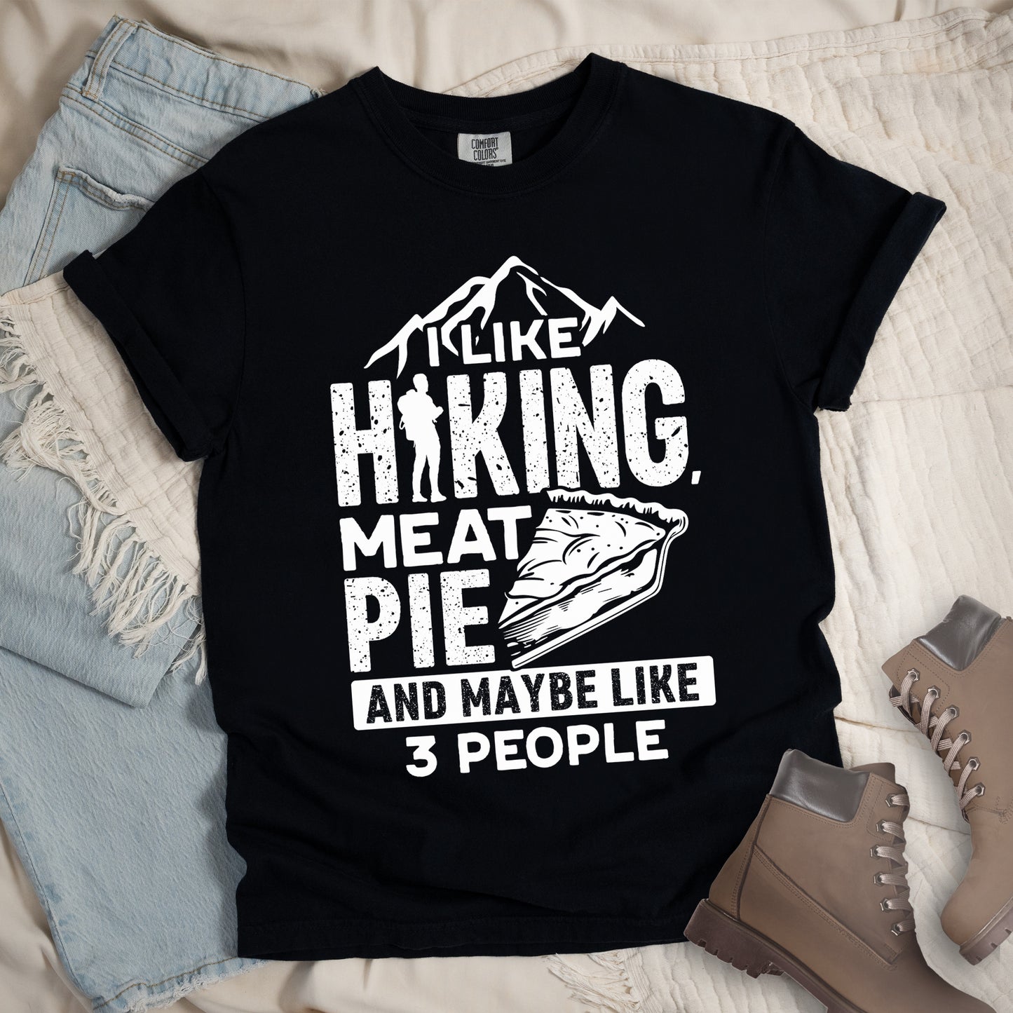Black t-shirt with white distressed text reading "I LIKE HIKING, MEAT PIE AND MAYBE LIKE 3 PEOPLE". Mountain silhouettes appear above "I LIKE". The letter "I" in "HIKING" is replaced with a hiker silhouette. Next to "MEAT PIE" is an illustration of a slice of pie. All text and graphics are in white with a weathered, vintage appearance. The design combines outdoor activities, food preferences, and introvert humor.