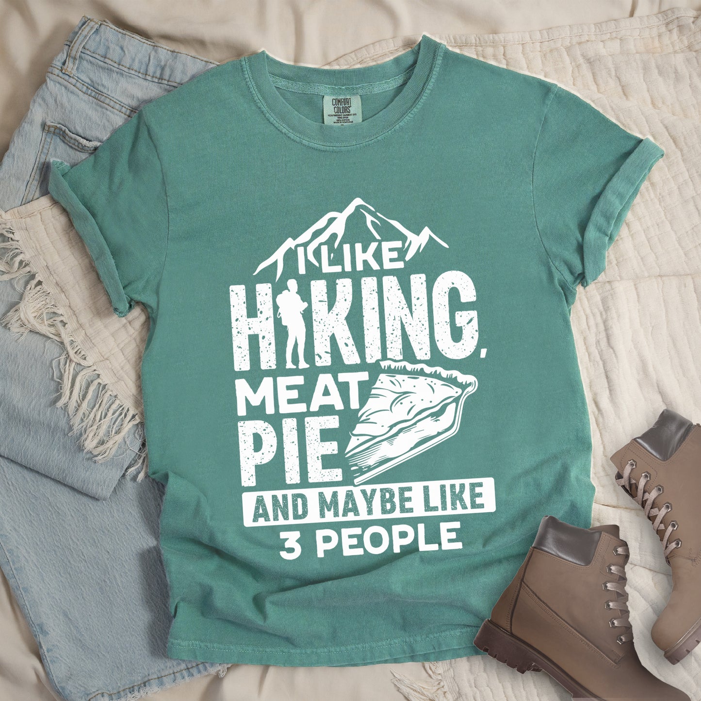Light Green t-shirt with white distressed text reading "I LIKE HIKING, MEAT PIE AND MAYBE LIKE 3 PEOPLE". Mountain silhouettes appear above "I LIKE". The letter "I" in "HIKING" is replaced with a hiker silhouette. Next to "MEAT PIE" is an illustration of a slice of pie. All text and graphics are in white with a weathered, vintage appearance. The design combines outdoor activities, food preferences, and introvert humor.