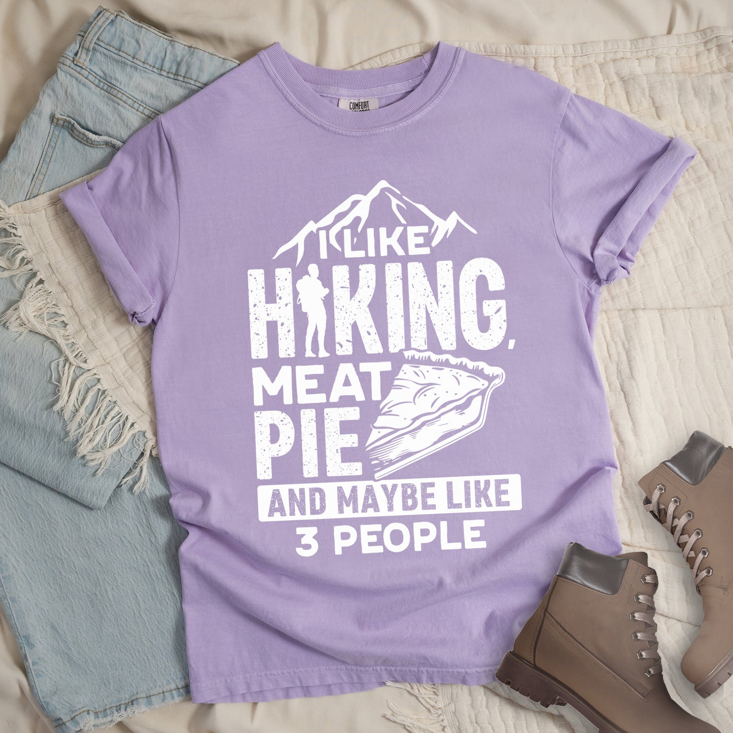 Orchid (Light Violet) t-shirt with white distressed text reading "I LIKE HIKING, MEAT PIE AND MAYBE LIKE 3 PEOPLE". Mountain silhouettes appear above "I LIKE". The letter "I" in "HIKING" is replaced with a hiker silhouette. Next to "MEAT PIE" is an illustration of a slice of pie. All text and graphics are in white with a weathered, vintage appearance. The design combines outdoor activities, food preferences, and introvert humor.