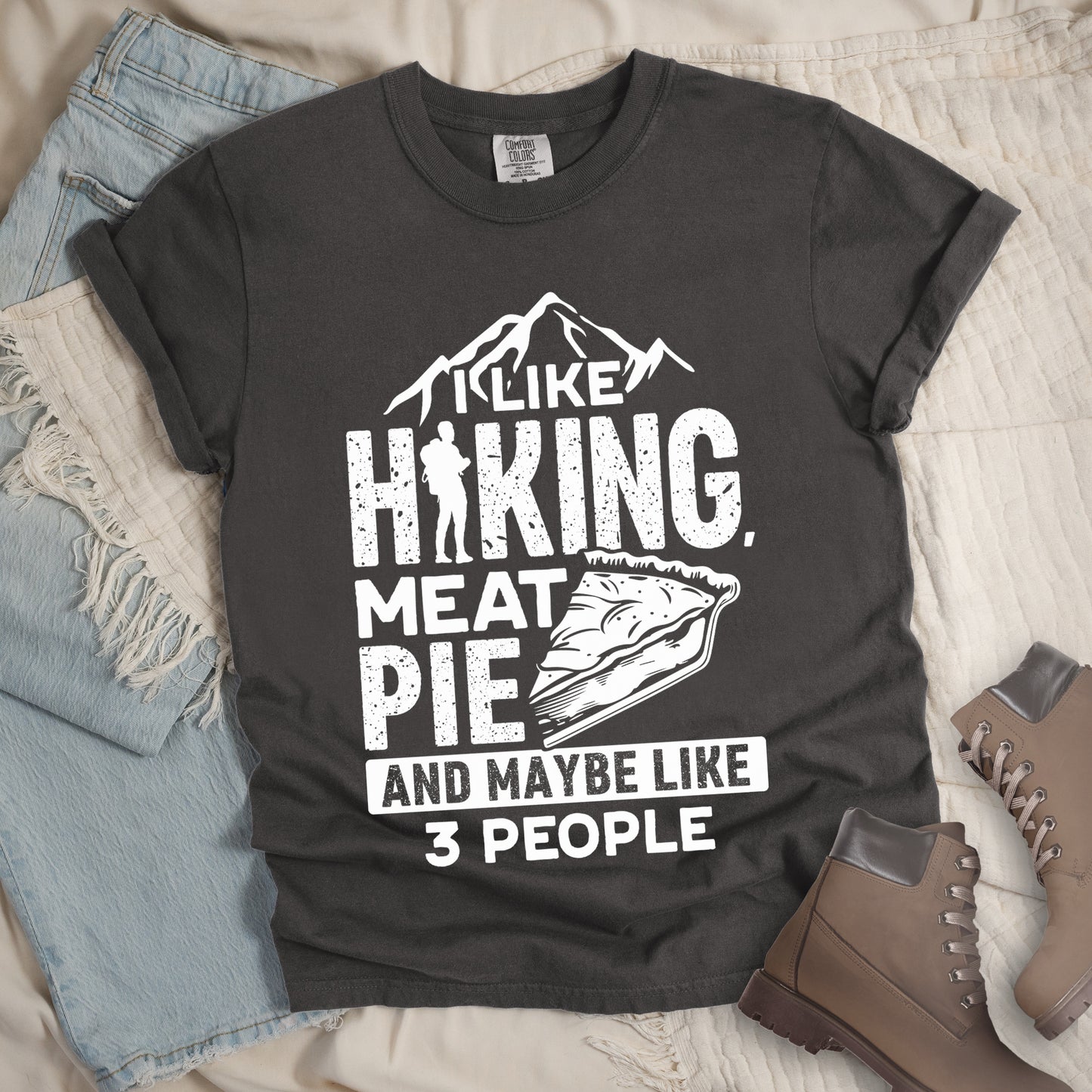 Pepper (Dark Gray) t-shirt with white distressed text reading "I LIKE HIKING, MEAT PIE AND MAYBE LIKE 3 PEOPLE". Mountain silhouettes appear above "I LIKE". The letter "I" in "HIKING" is replaced with a hiker silhouette. Next to "MEAT PIE" is an illustration of a slice of pie. All text and graphics are in white with a weathered, vintage appearance. The design combines outdoor activities, food preferences, and introvert humor.