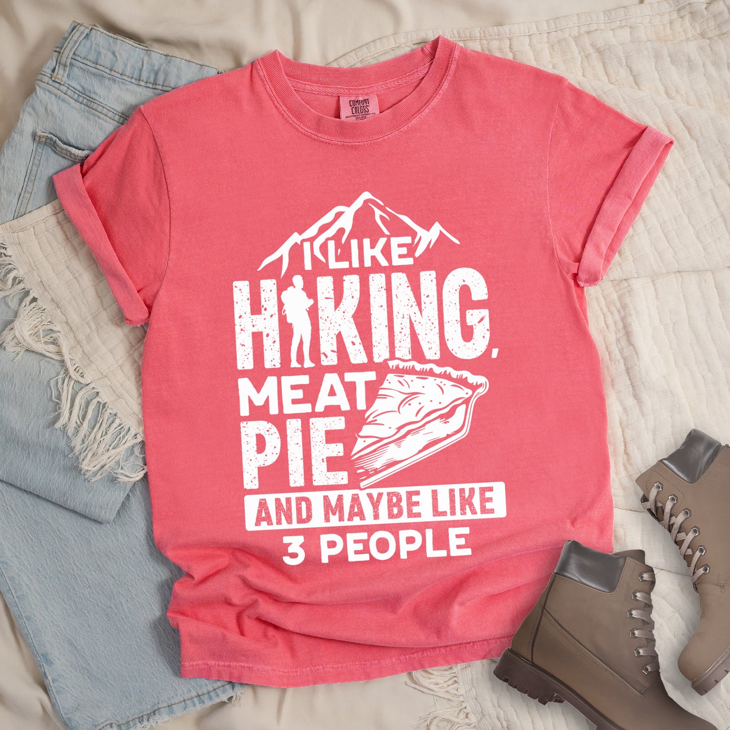 Watermelon (Light Coral) t-shirt with white distressed text reading "I LIKE HIKING, MEAT PIE AND MAYBE LIKE 3 PEOPLE". Mountain silhouettes appear above "I LIKE". The letter "I" in "HIKING" is replaced with a hiker silhouette. Next to "MEAT PIE" is an illustration of a slice of pie. All text and graphics are in white with a weathered, vintage appearance. The design combines outdoor activities, food preferences, and introvert humor.
