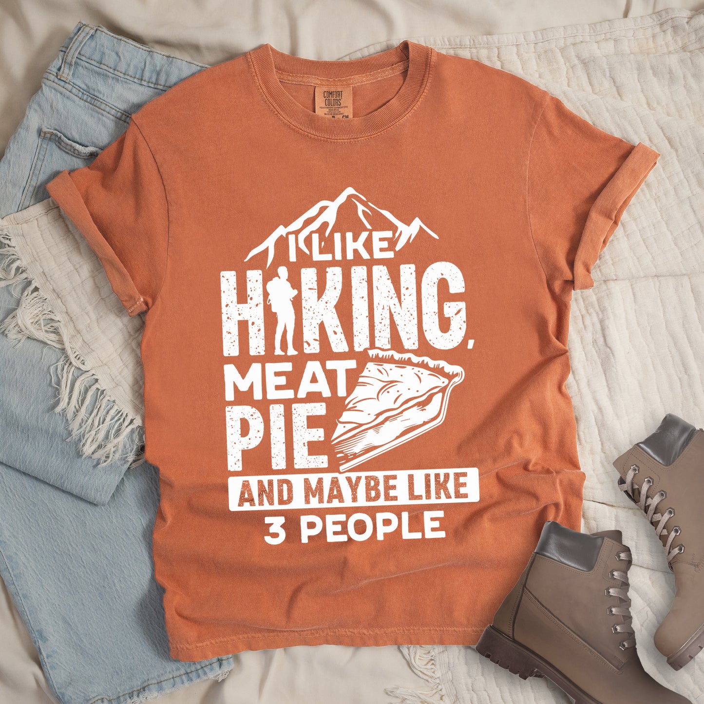 Yam (Brown) t-shirt with white distressed text reading "I LIKE HIKING, MEAT PIE AND MAYBE LIKE 3 PEOPLE". Mountain silhouettes appear above "I LIKE". The letter "I" in "HIKING" is replaced with a hiker silhouette. Next to "MEAT PIE" is an illustration of a slice of pie. All text and graphics are in white with a weathered, vintage appearance. The design combines outdoor activities, food preferences, and introvert humor.