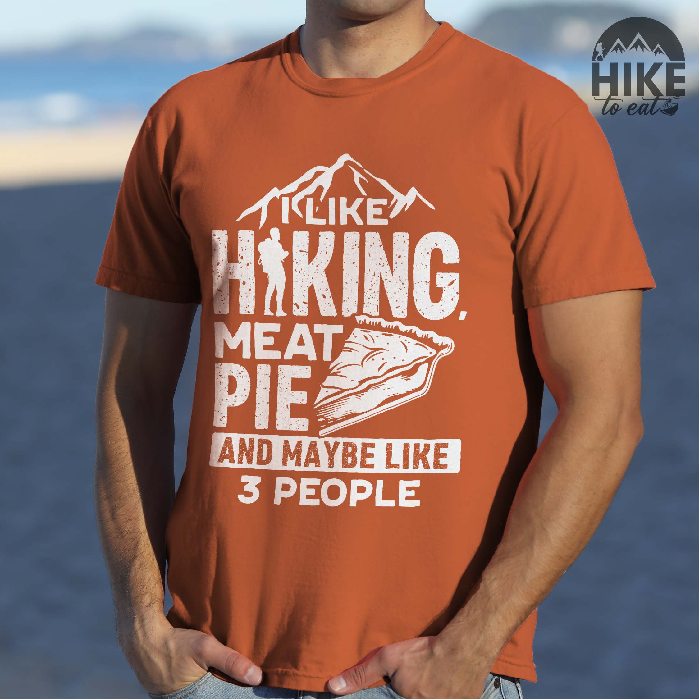 Man wearing yam "I like hiking, meat pie and maybe like 3 people" colored vintage tee with bold typography, mountains, birds, illustrated slice of meat pie, and backpacker for pie lovers.