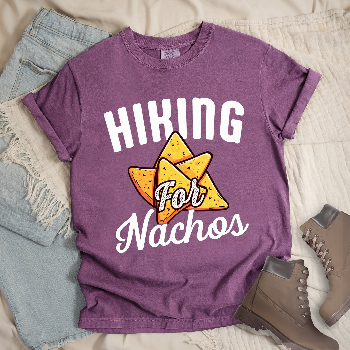 Berry (Dark Violet) t-shirt with white text reading "HIKING For Nachos". "HIKING" appears in large white letters at the top, "For" in white script across the middle, and "Nachos" in white script at the bottom. The centerpiece of the design is an illustration of tortilla chips arranged in a star-like pattern, colored yellow-orange with flecks to represent seasonings. The design connects hiking activity with food motivation.