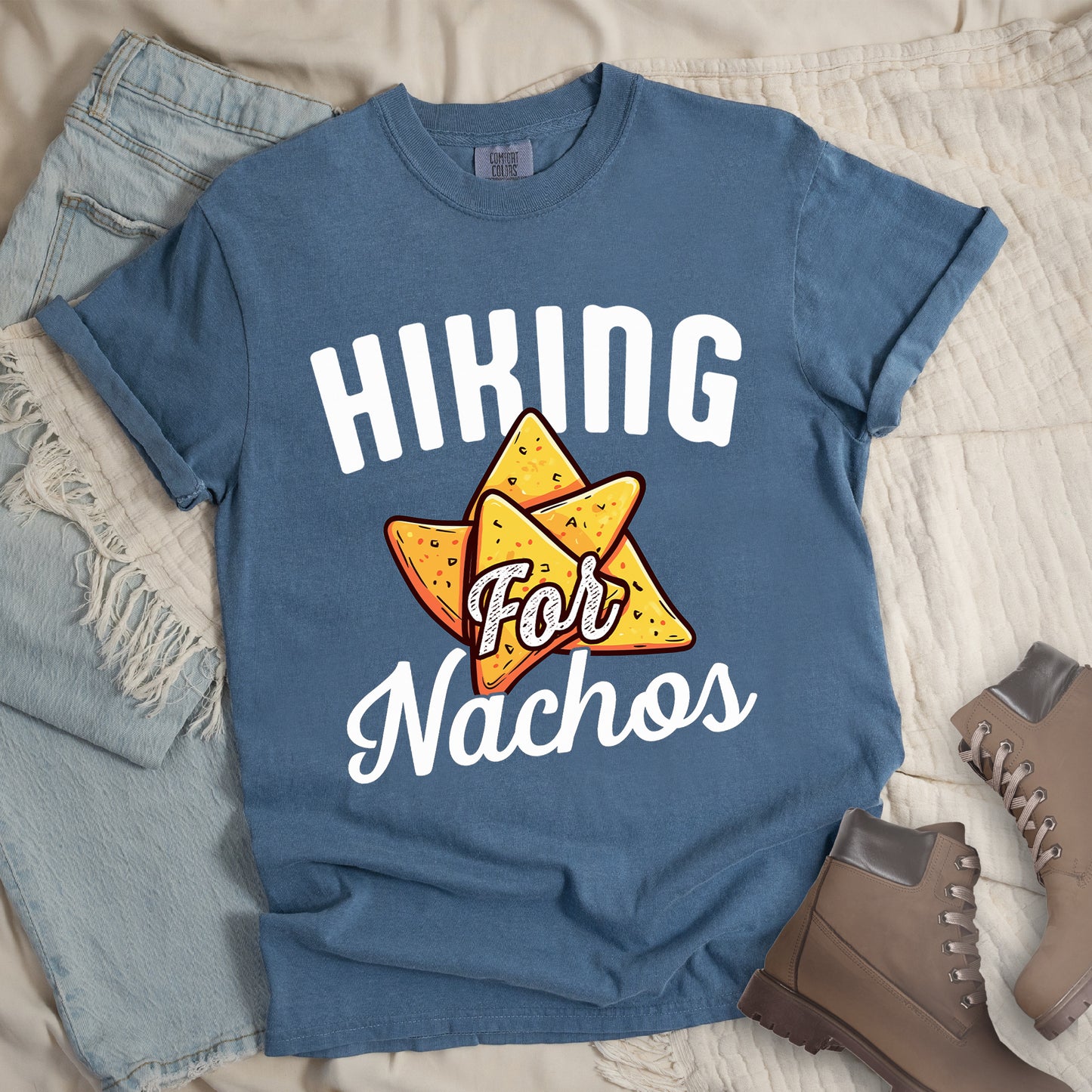 Blue Jean (Denim Blue) t-shirt with white text reading "HIKING For Nachos". "HIKING" appears in large white letters at the top, "For" in white script across the middle, and "Nachos" in white script at the bottom. The centerpiece of the design is an illustration of tortilla chips arranged in a star-like pattern, colored yellow-orange with flecks to represent seasonings. The design connects hiking activity with food motivation.