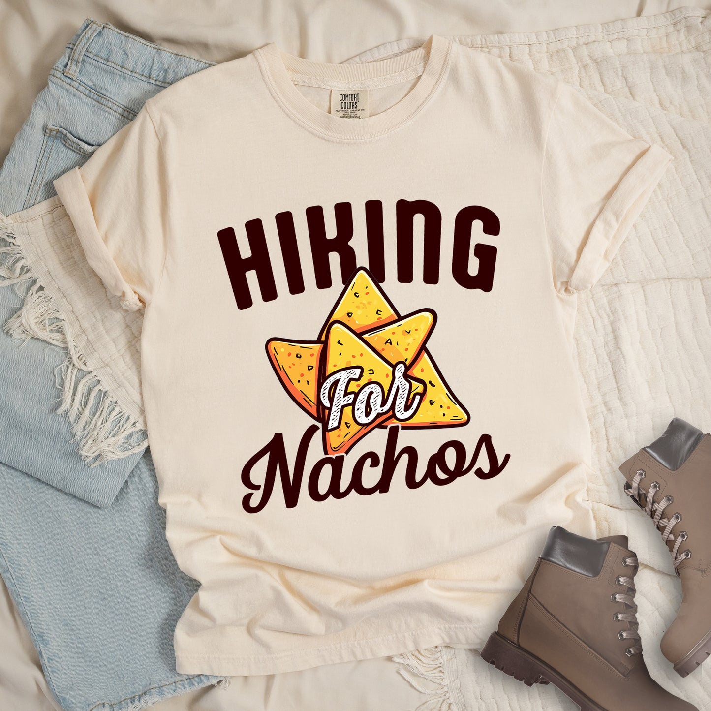 Ivory (Cream White) t-shirt with deep brown text reading "HIKING For Nachos". "HIKING" appears in large deep brown letters at the top, "For" in white script across the middle, and "Nachos" in deep brown script at the bottom. The centerpiece of the design is an illustration of tortilla chips arranged in a star-like pattern, colored yellow-orange with flecks to represent seasonings. The design connects hiking activity with food motivation.