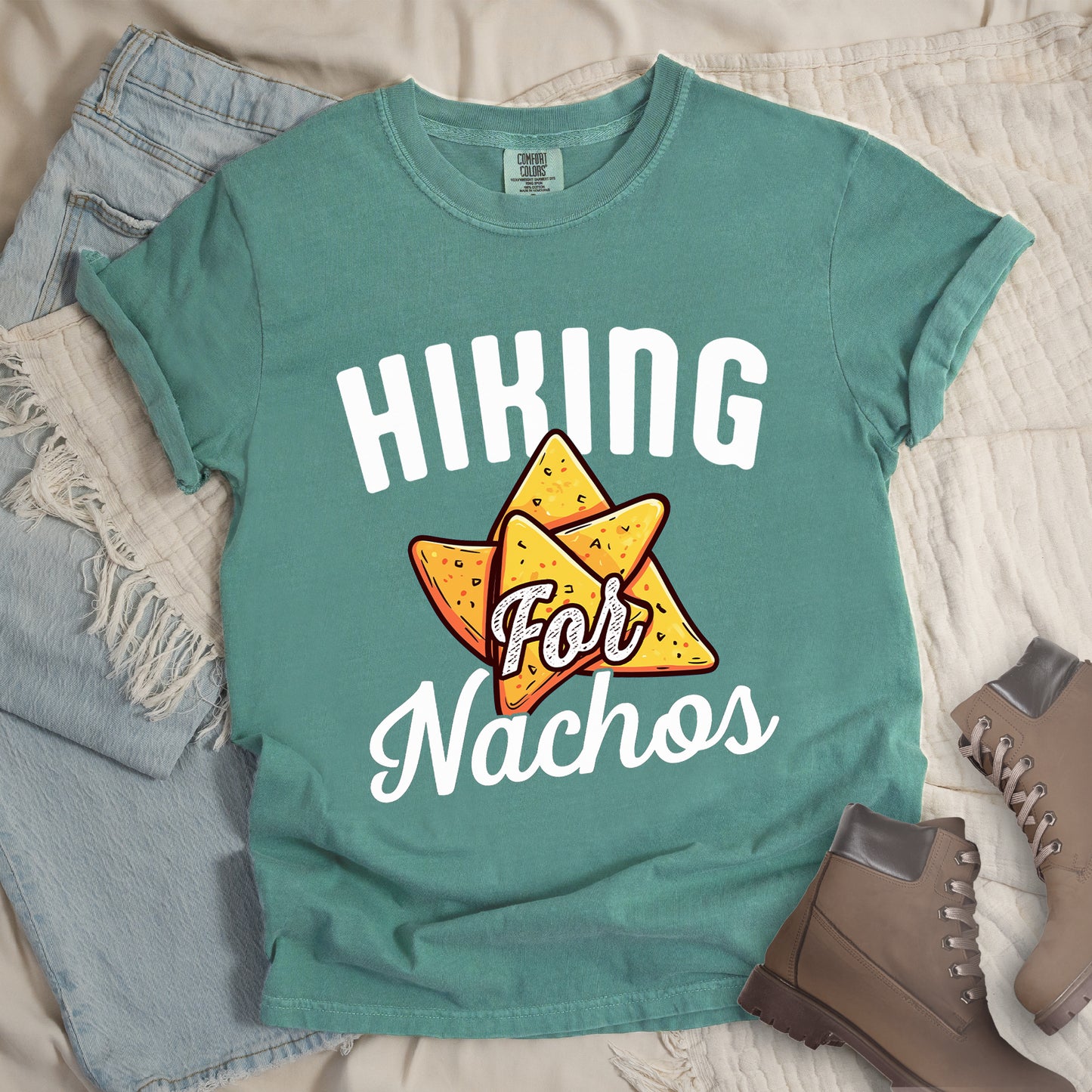 Light Green t-shirt with white text reading "HIKING For Nachos". "HIKING" appears in large white letters at the top, "For" in white script across the middle, and "Nachos" in white script at the bottom. The centerpiece of the design is an illustration of tortilla chips arranged in a star-like pattern, colored yellow-orange with flecks to represent seasonings. The design connects hiking activity with food motivation.