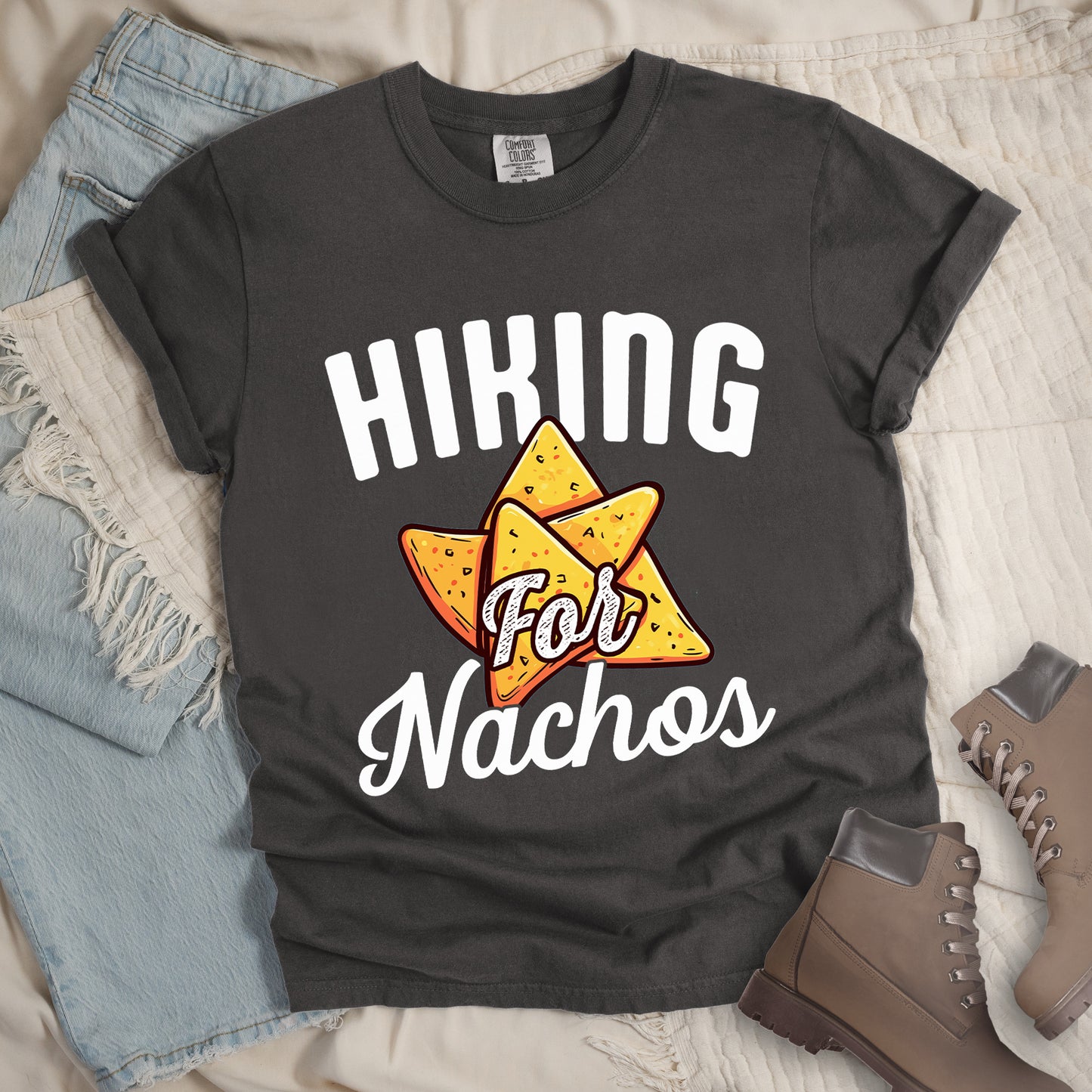 Pepper (Dark Gray) t-shirt with white text reading "HIKING For Nachos". "HIKING" appears in large white letters at the top, "For" in white script across the middle, and "Nachos" in white script at the bottom. The centerpiece of the design is an illustration of tortilla chips arranged in a star-like pattern, colored yellow-orange with flecks to represent seasonings. The design connects hiking activity with food motivation.