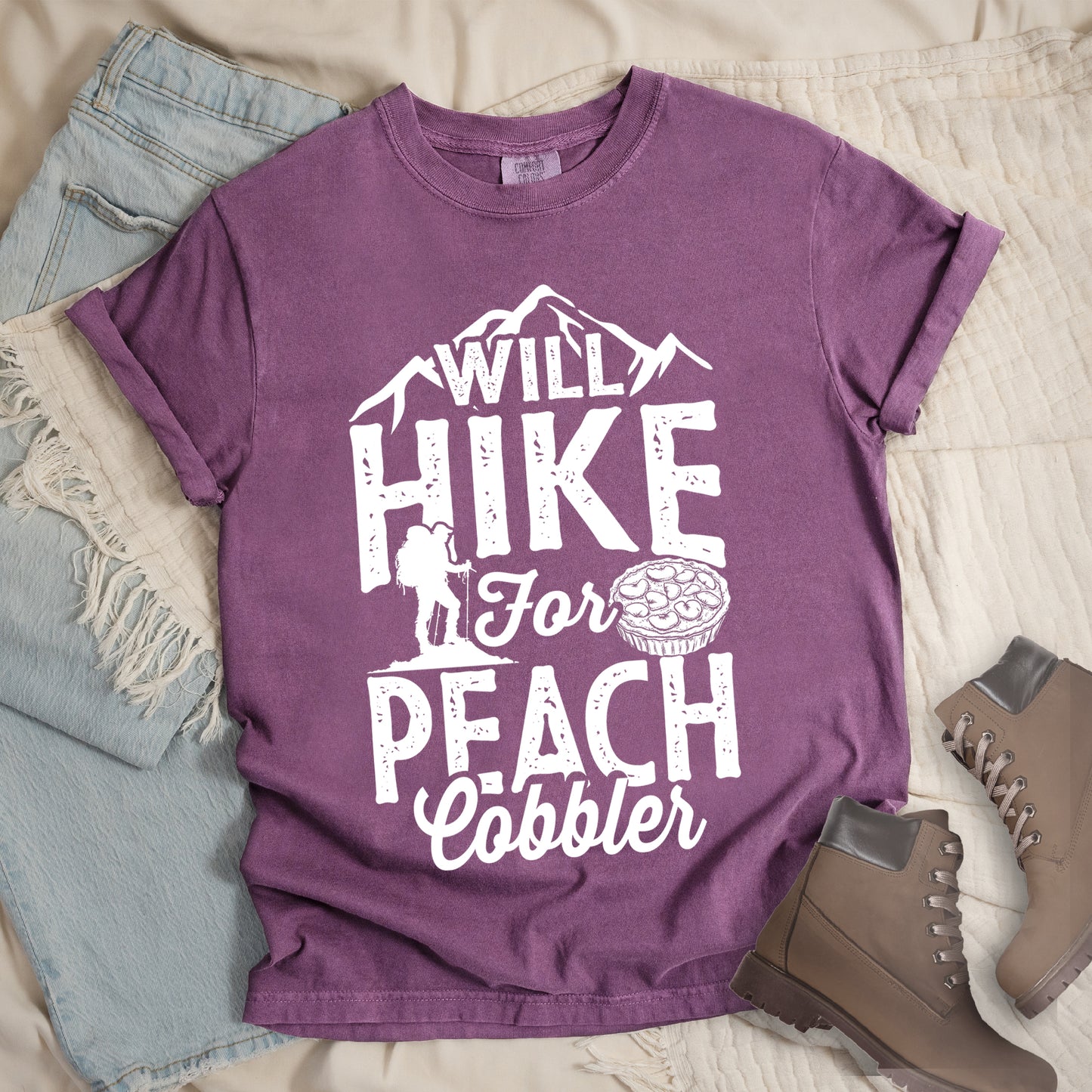 Berry (Dark Violet) t-shirt with white text reading "WILL HIKE FOR PEACH COBBLER". "WILL HIKE" appears in large distressed letters at the top with mountain silhouettes, "For" in white script across the middle, and "PEACH COBBLER" in large distressed letters at the bottom. The design includes a small hiker figure with backpack on the left and an illustration of a peach cobbler pie. The design humorously connects hiking activity with dessert motivation.