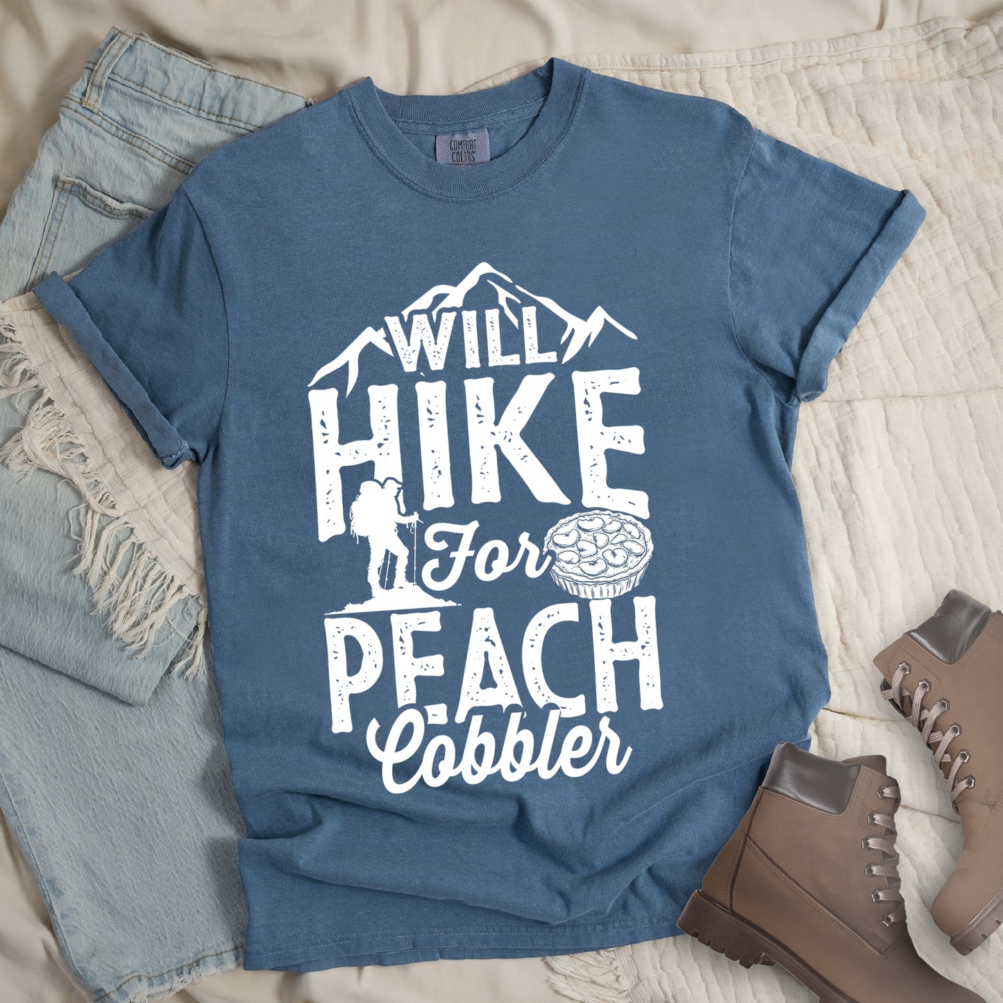 Blue Jean (Denim Blue) t-shirt with white text reading "WILL HIKE FOR PEACH COBBLER". "WILL HIKE" appears in large distressed letters at the top with mountain silhouettes, "For" in white script across the middle, and "PEACH COBBLER" in large distressed letters at the bottom. The design includes a small hiker figure with backpack on the left and an illustration of a peach cobbler pie. The design humorously connects hiking activity with dessert motivation.