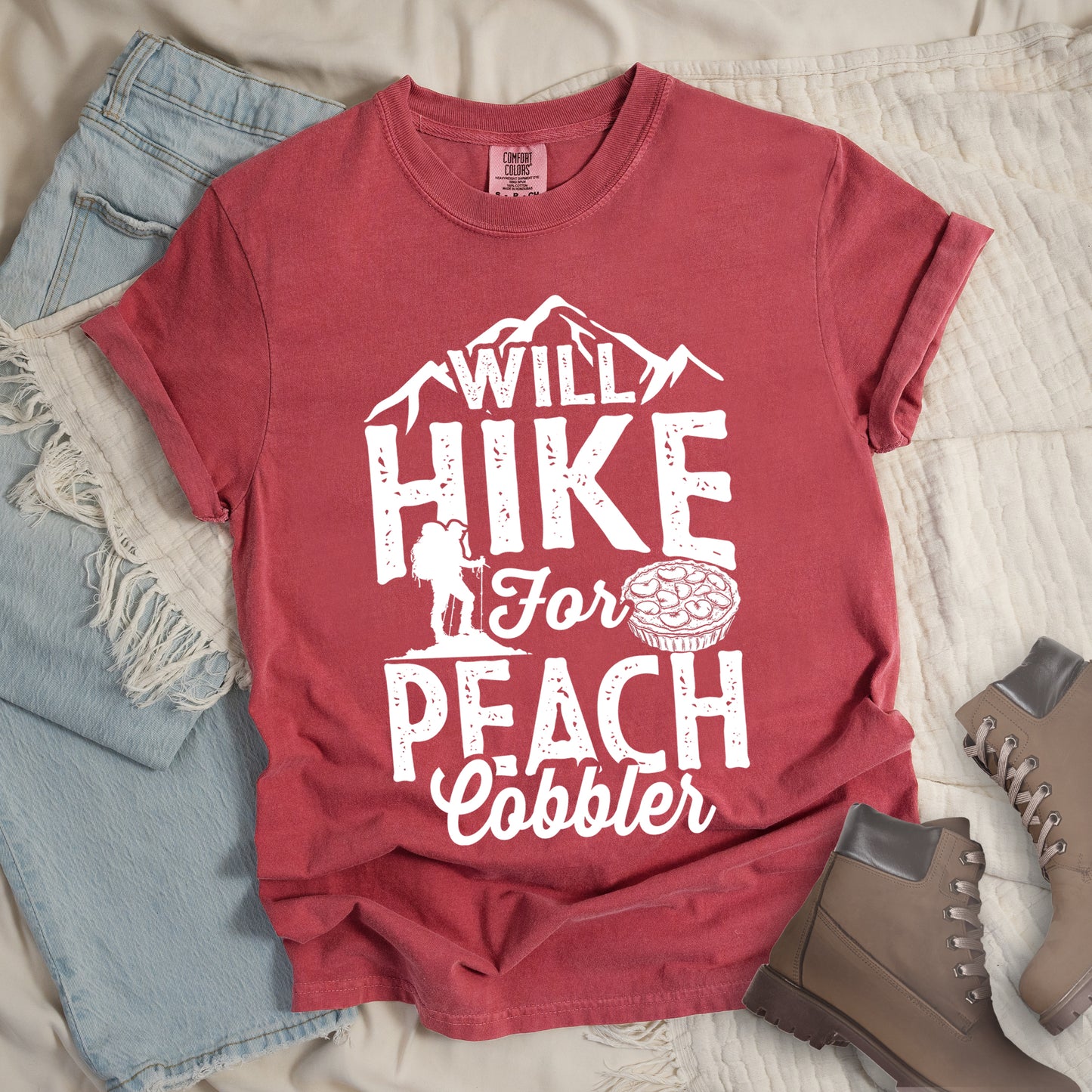 Crimson (Dark Pink) t-shirt with white text reading "WILL HIKE FOR PEACH COBBLER". "WILL HIKE" appears in large distressed letters at the top with mountain silhouettes, "For" in white script across the middle, and "PEACH COBBLER" in large distressed letters at the bottom. The design includes a small hiker figure with backpack on the left and an illustration of a peach cobbler pie. The design humorously connects hiking activity with dessert motivation.