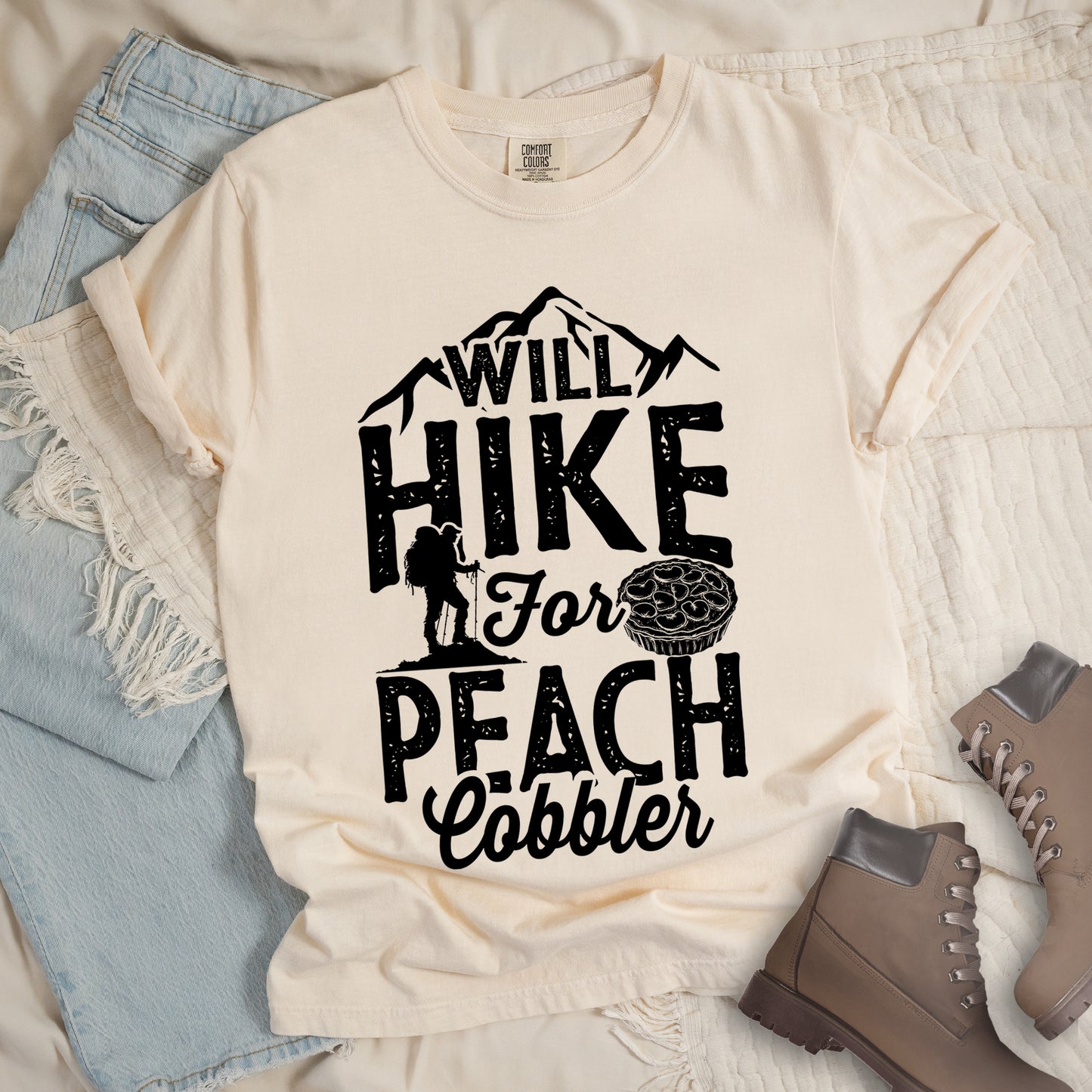 Ivory (Cream White) t-shirt with black text reading "WILL HIKE FOR PEACH COBBLER". "WILL HIKE" appears in large distressed letters at the top with mountain silhouettes, "For" in black script across the middle, and "PEACH COBBLER" in large distressed letters at the bottom. The design includes a small hiker figure with backpack on the left and an illustration of a peach cobbler pie. The design humorously connects hiking activity with dessert motivation.