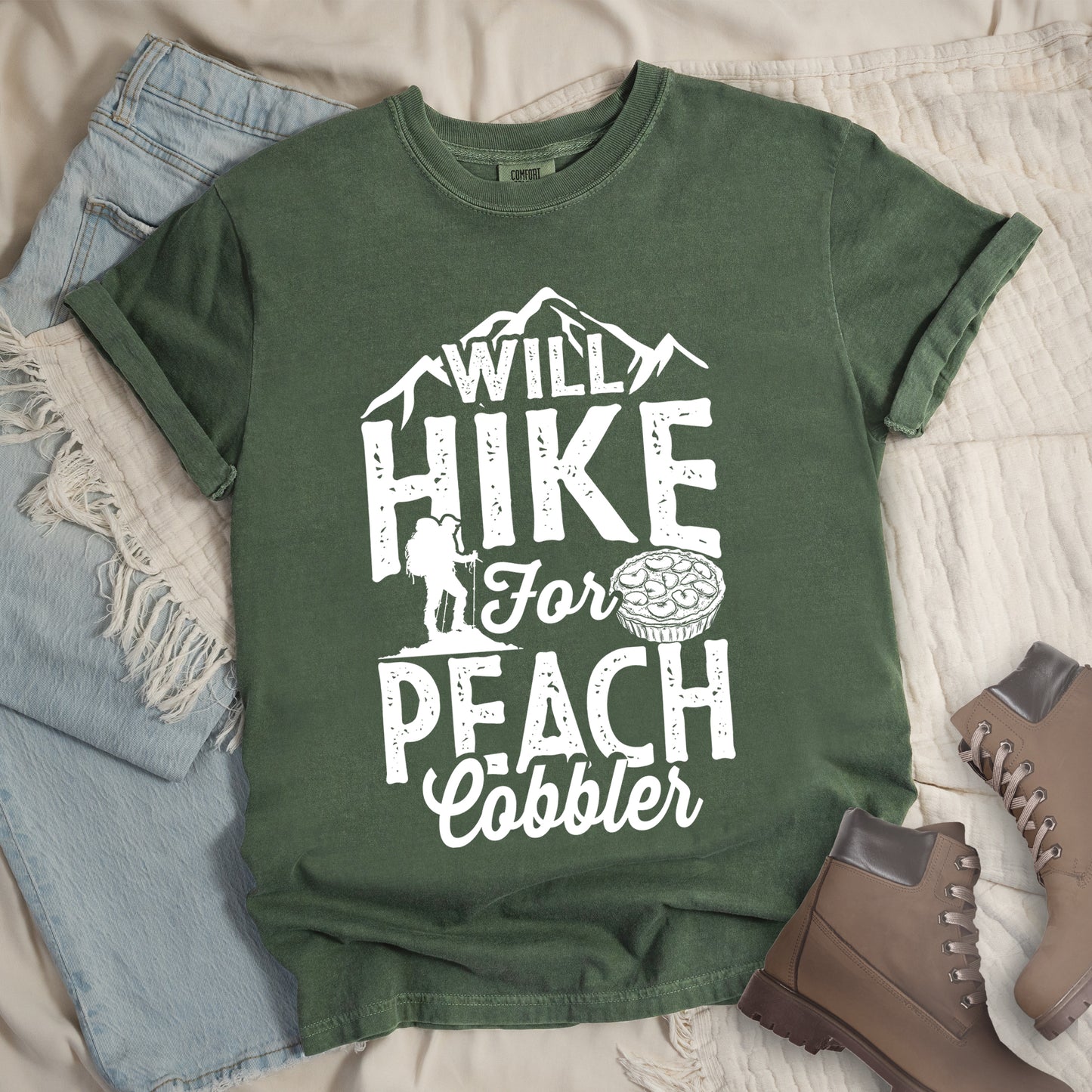 Moss (Dark Green) t-shirt with white text reading "WILL HIKE FOR PEACH COBBLER". "WILL HIKE" appears in large distressed letters at the top with mountain silhouettes, "For" in white script across the middle, and "PEACH COBBLER" in large distressed letters at the bottom. The design includes a small hiker figure with backpack on the left and an illustration of a peach cobbler pie. The design humorously connects hiking activity with dessert motivation.