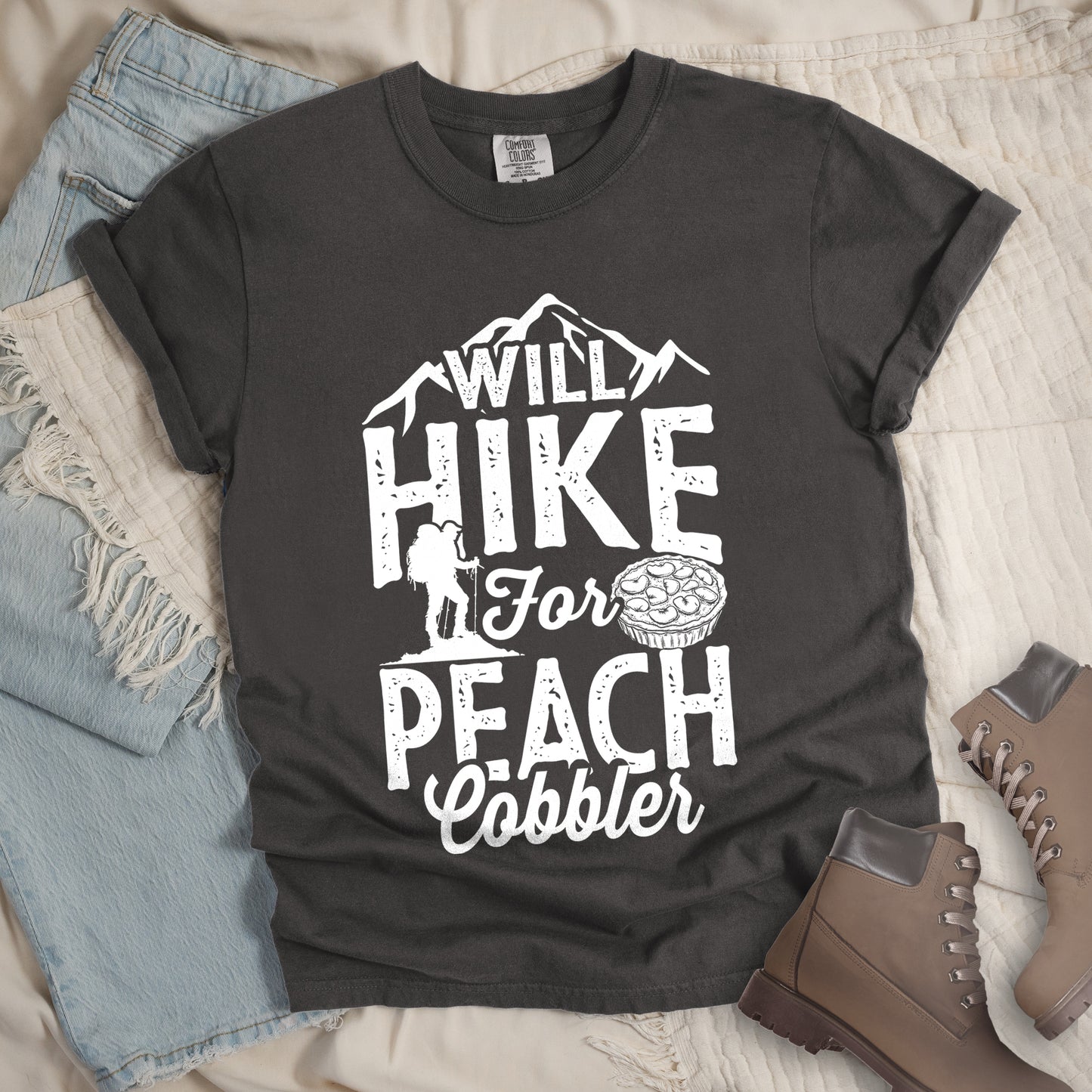Pepper (Dark Gray) t-shirt with white text reading "WILL HIKE FOR PEACH COBBLER". "WILL HIKE" appears in large distressed letters at the top with mountain silhouettes, "For" in white script across the middle, and "PEACH COBBLER" in large distressed letters at the bottom. The design includes a small hiker figure with backpack on the left and an illustration of a peach cobbler pie. The design humorously connects hiking activity with dessert motivation.