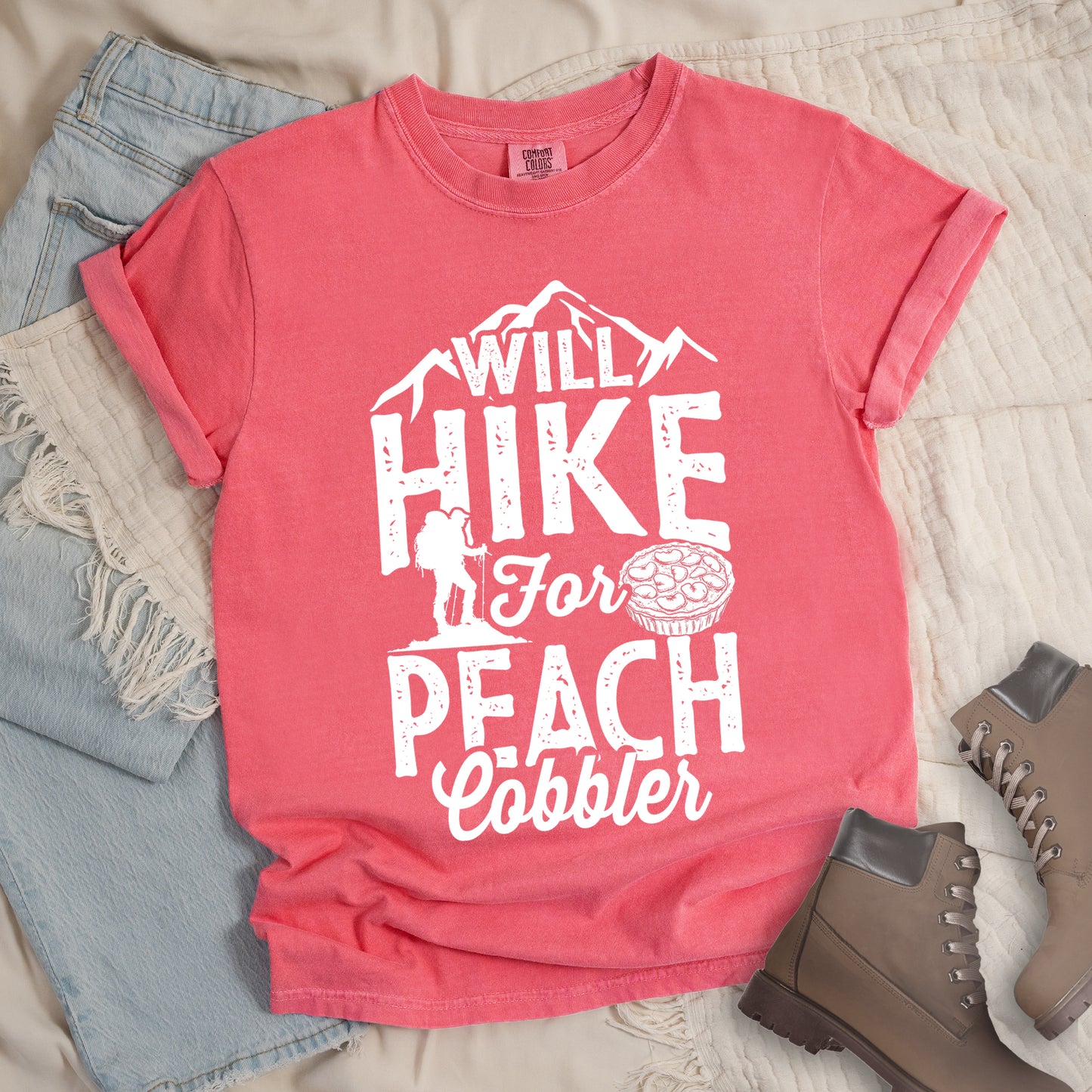 Watermelon (Light Coral) t-shirt with white text reading "WILL HIKE FOR PEACH COBBLER". "WILL HIKE" appears in large distressed letters at the top with mountain silhouettes, "For" in white script across the middle, and "PEACH COBBLER" in large distressed letters at the bottom. The design includes a small hiker figure with backpack on the left and an illustration of a peach cobbler pie. The design humorously connects hiking activity with dessert motivation.