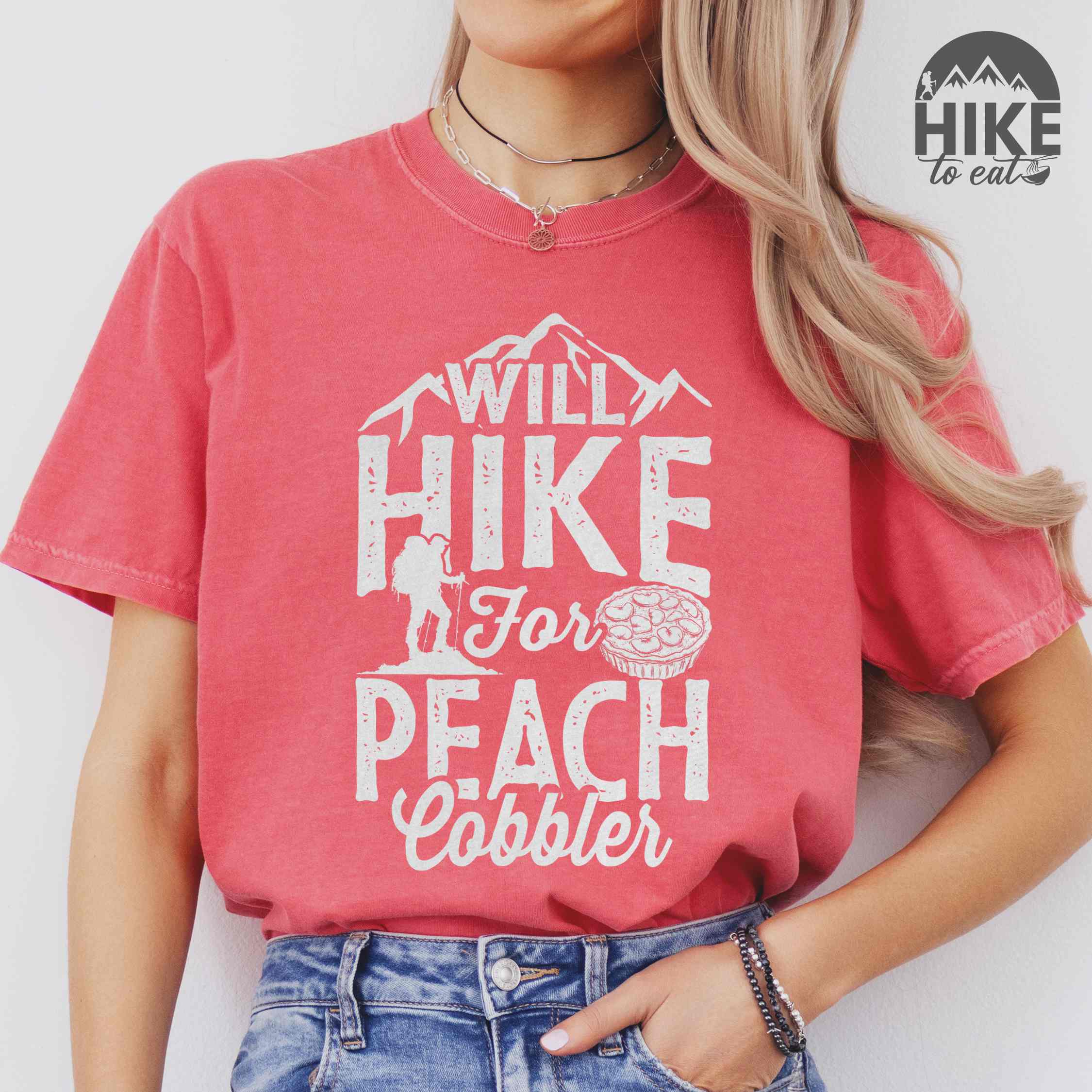 Woman wearing watermelon "Will hike for peach cobbler" colored vintage tee with bold typography, mountain, birds, illustrated peach cobbler, and backpacker looking for sweet treats.