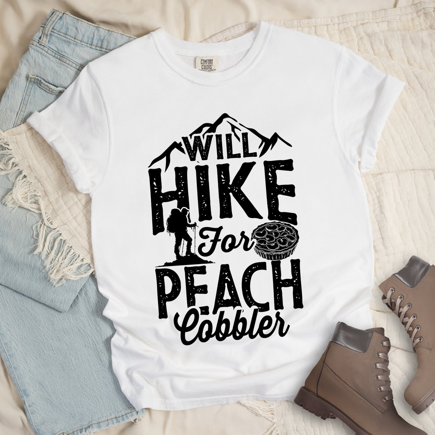 White t-shirt with black text reading "WILL HIKE FOR PEACH COBBLER". "WILL HIKE" appears in large distressed letters at the top with mountain silhouettes, "For" in black script across the middle, and "PEACH COBBLER" in large distressed letters at the bottom. The design includes a small hiker figure with backpack on the left and an illustration of a peach cobbler pie. The design humorously connects hiking activity with dessert motivation.