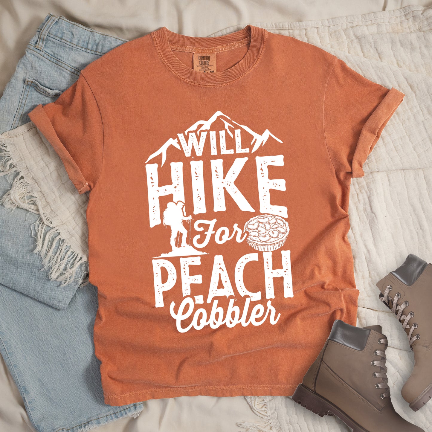 Yam (Brown) t-shirt with white text reading "WILL HIKE FOR PEACH COBBLER". "WILL HIKE" appears in large distressed letters at the top with mountain silhouettes, "For" in white script across the middle, and "PEACH COBBLER" in large distressed letters at the bottom. The design includes a small hiker figure with backpack on the left and an illustration of a peach cobbler pie. The design humorously connects hiking activity with dessert motivation.