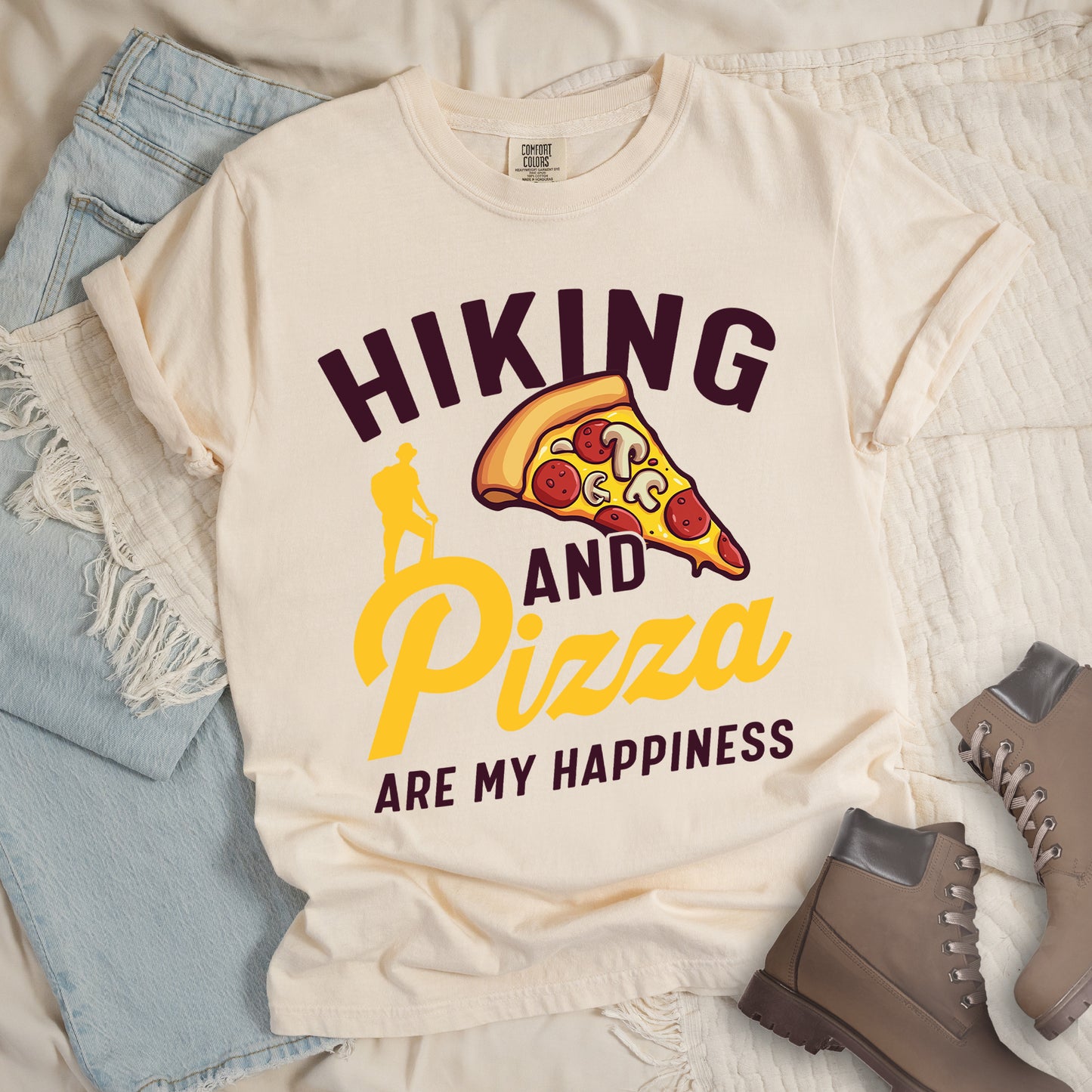 Ivory (Cream White) t-shirt with deep brown and yellow text reading "HIKING AND Pizza ARE MY HAPPINESS". "HIKING" appears in deep brown at the top, "AND" in deep brown in the middle, "Pizza" in yellow script, and "ARE MY HAPPINESS" in deep brown at the bottom. Features a colorful pizza slice with pepperoni and mushrooms in the center-right and a yellow silhouette of a hiker on the left side above "AND Pizza ARE MY HAPPINESS". The design combines outdoor activity with food enjoyment.