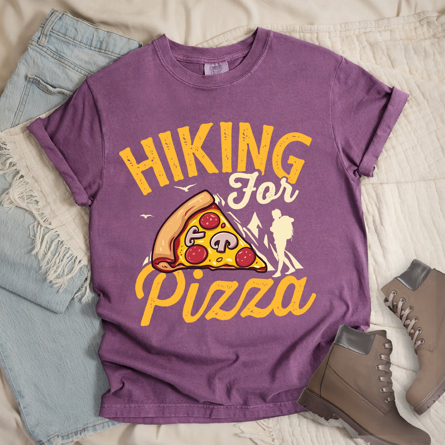 Berry (Dark Violet) t-shirt with yellow distressed text reading "HIKING For Pizza". "HIKING" appears in large yellow letters at the top, "For" in cream script in the middle, and "Pizza" in yellow script at the bottom. Features a detailed pizza slice with pepperoni and mushrooms in the center and a cream silhouette of a hiker on the right side against mountain outlines. Small bird silhouettes appear near the mountains. The design connects hiking activity with food reward.