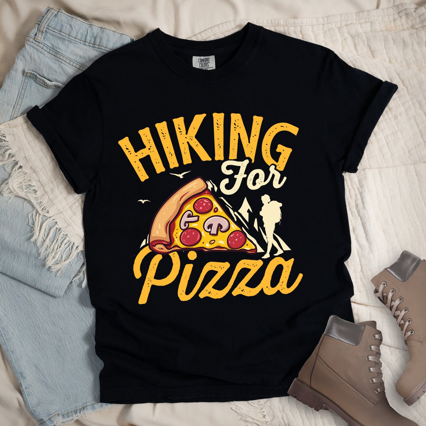 Black t-shirt with yellow distressed text reading "HIKING For Pizza". "HIKING" appears in large yellow letters at the top, "For" in cream script in the middle, and "Pizza" in yellow script at the bottom. Features a detailed pizza slice with pepperoni and mushrooms in the center and a cream silhouette of a hiker on the right side against mountain outlines. Small bird silhouettes appear near the mountains. The design connects hiking activity with food reward.