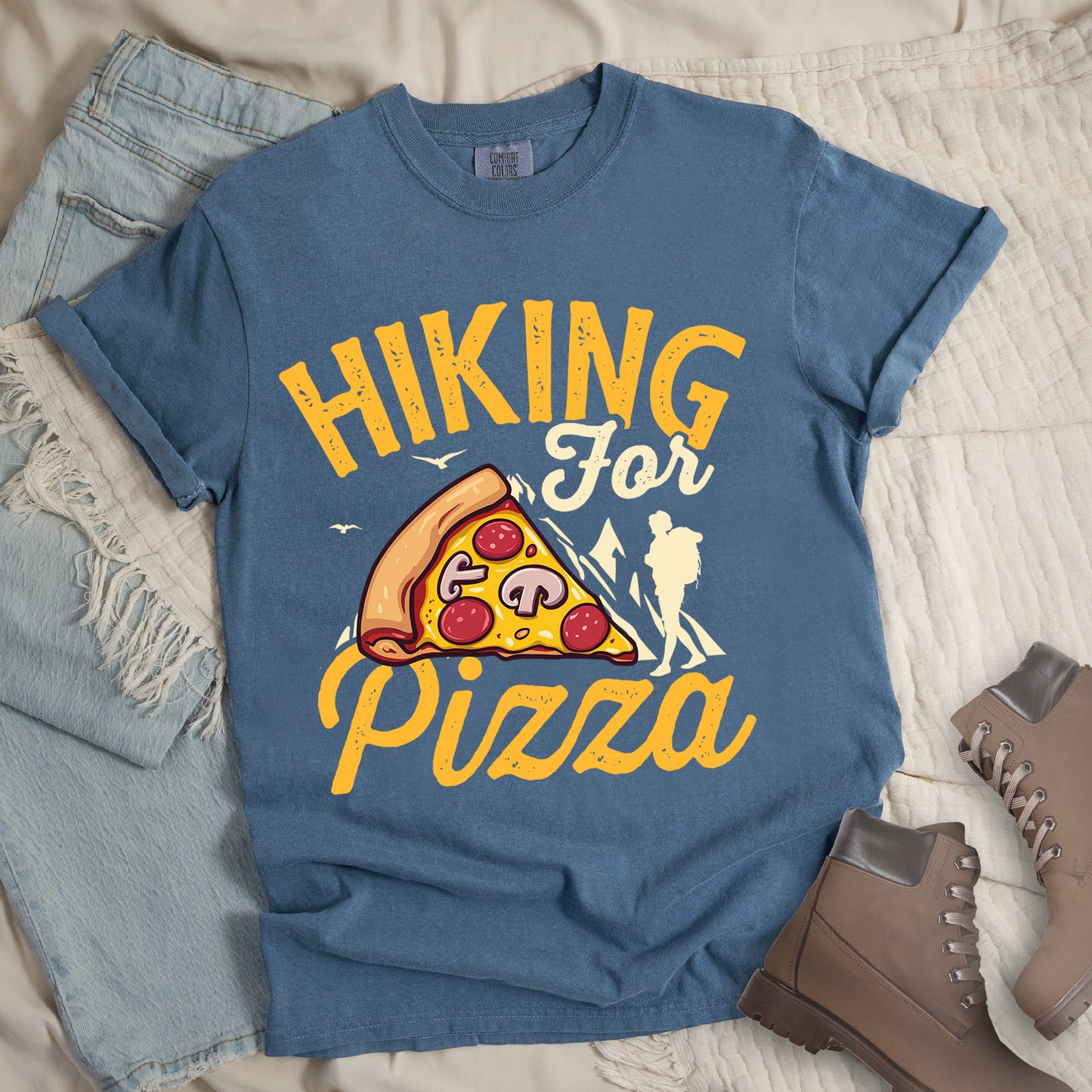 Blue Jean (Denim Blue) t-shirt with yellow distressed text reading "HIKING For Pizza". "HIKING" appears in large yellow letters at the top, "For" in cream script in the middle, and "Pizza" in yellow script at the bottom. Features a detailed pizza slice with pepperoni and mushrooms in the center and a cream silhouette of a hiker on the right side against mountain outlines. Small bird silhouettes appear near the mountains. The design connects hiking activity with food reward.