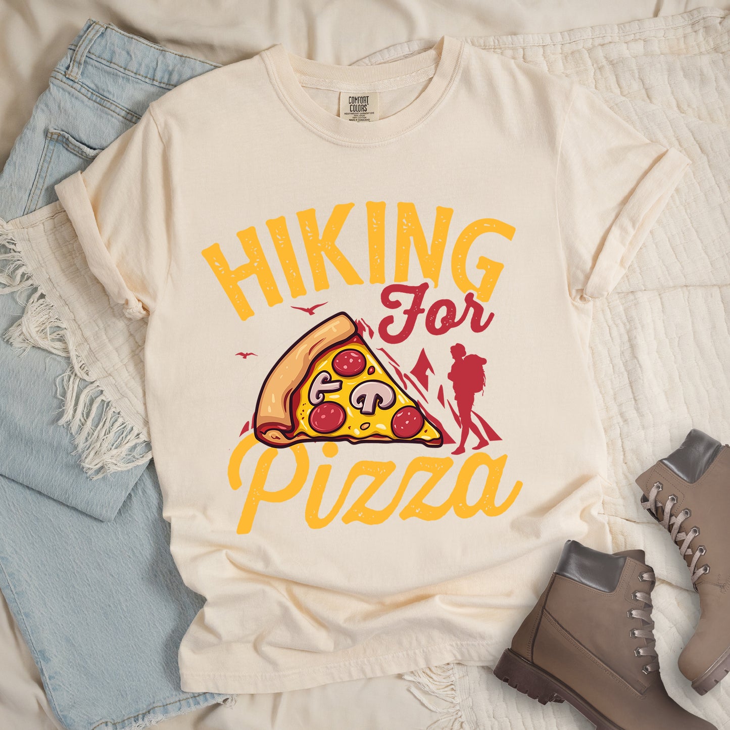 Ivory (Cream White) t-shirt with yellow distressed text reading "HIKING For Pizza". "HIKING" appears in large yellow letters at the top, "For" in red script in the middle, and "Pizza" in yellow script at the bottom. Features a detailed pizza slice with pepperoni and mushrooms in the center and a red silhouette of a hiker on the right side against mountain outlines. Small bird silhouettes appear near the mountains. The design connects hiking activity with food reward.
