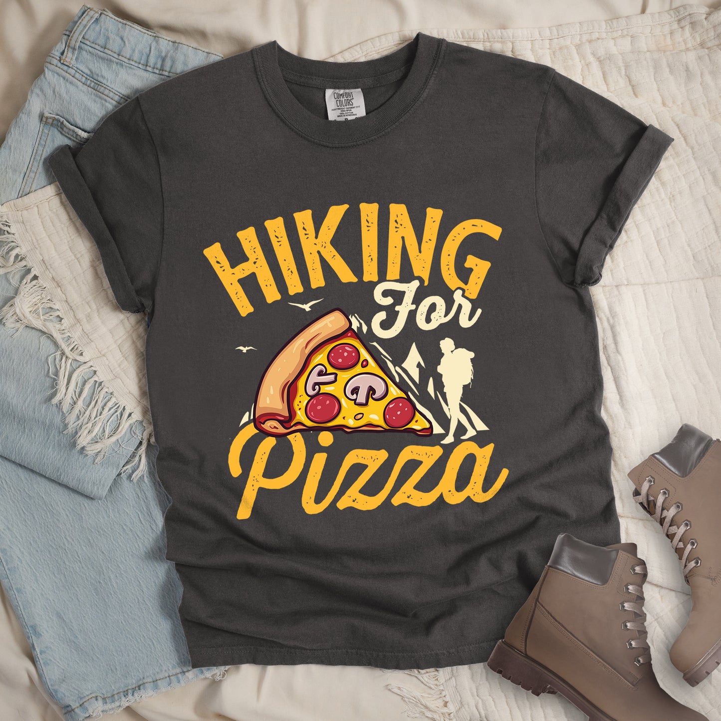 Pepper (Dark Gray) t-shirt with yellow distressed text reading "HIKING For Pizza". "HIKING" appears in large yellow letters at the top, "For" in cream script in the middle, and "Pizza" in yellow script at the bottom. Features a detailed pizza slice with pepperoni and mushrooms in the center and a cream silhouette of a hiker on the right side against mountain outlines. Small bird silhouettes appear near the mountains. The design connects hiking activity with food reward.