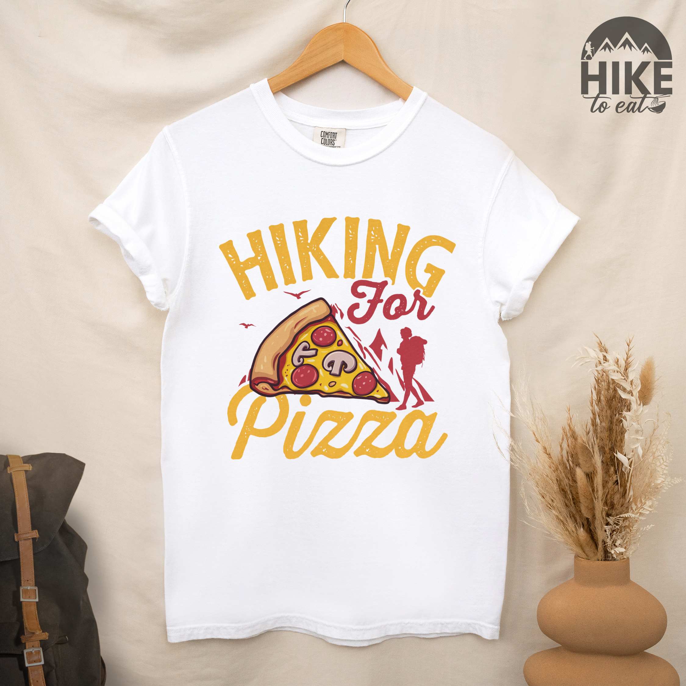 Hanging white "Hiking for Pizza" colored vintage tee with bold typography, birds, mountains, illustrated pizza slice, and backpacker for pizza lovers.