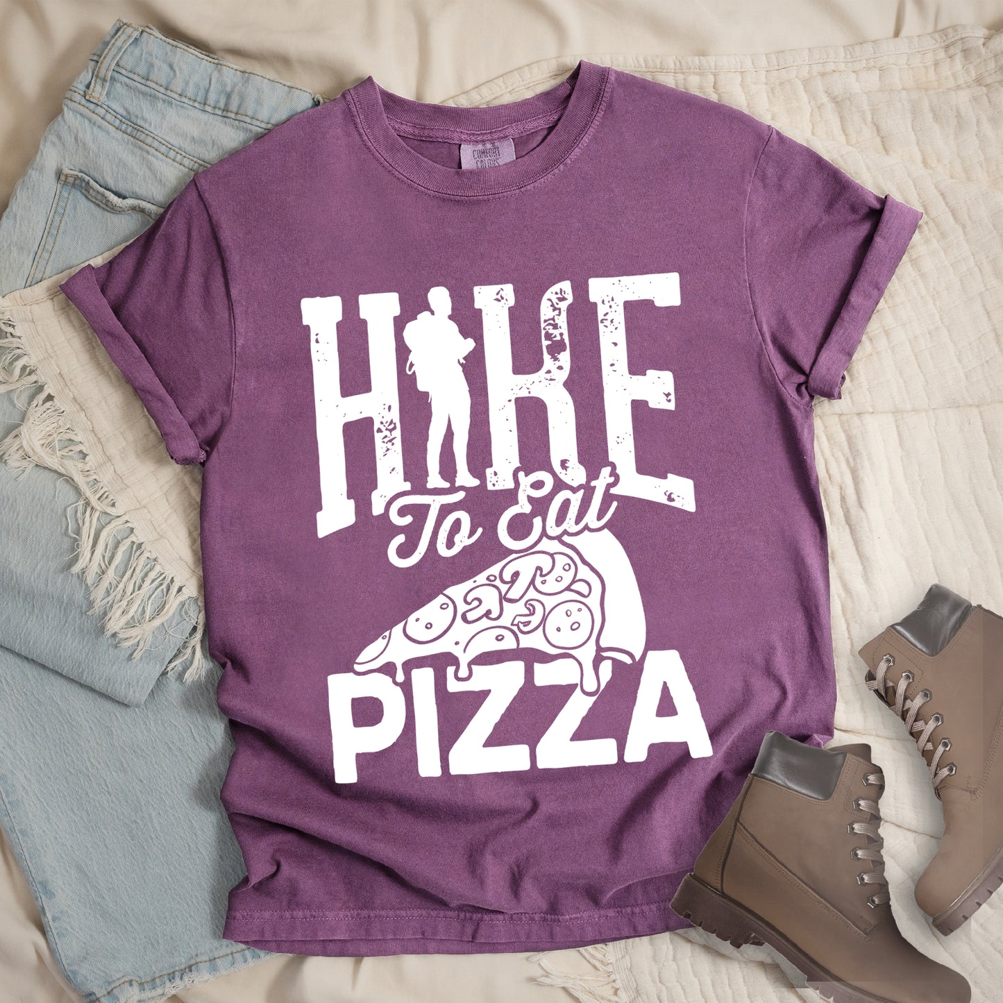 Berry (Dark Violet) t-shirt with white distressed text reading "HIKE To Eat PIZZA". "HIKE" appears in large block letters at the top with a silhouette of a hiker with backpack integrated into the "I", "To Eat" in script in the middle, and "PIZZA" in large block letters at the bottom. Features a simple line drawing of a pizza slice with toppings between the phrases. The design humorously connects hiking activity with pizza as a reward.
