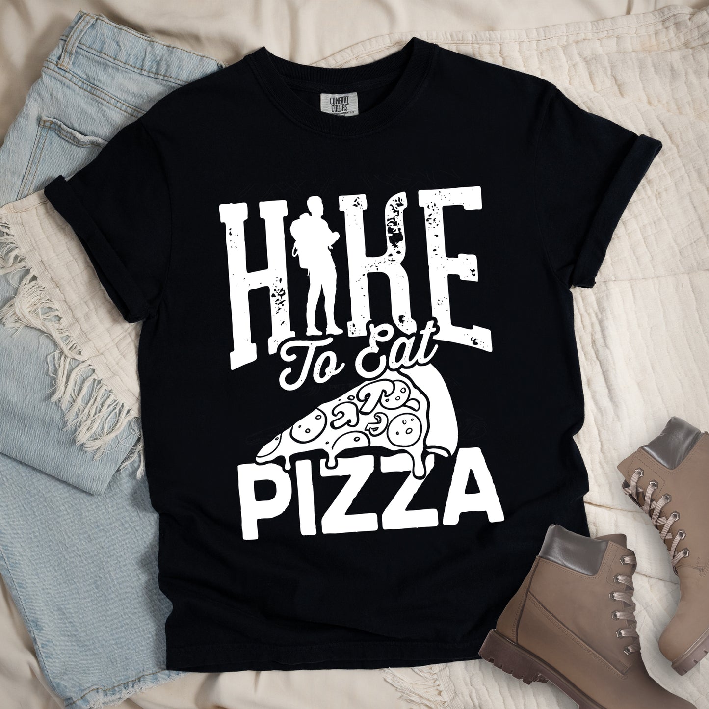 Black t-shirt with white distressed text reading "HIKE To Eat PIZZA". "HIKE" appears in large block letters at the top with a silhouette of a hiker with backpack integrated into the "I", "To Eat" in script in the middle, and "PIZZA" in large block letters at the bottom. Features a simple line drawing of a pizza slice with toppings between the phrases. The design humorously connects hiking activity with pizza as a reward.