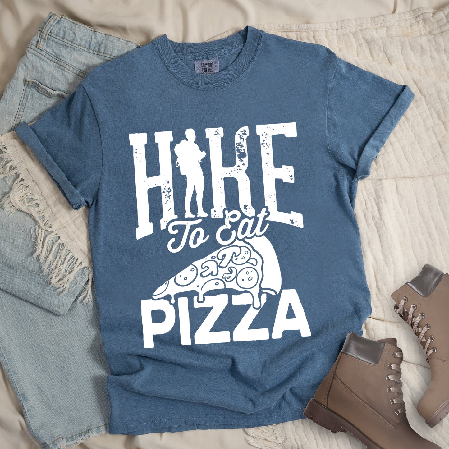 Blue Jean (Denim Blue) t-shirt with white distressed text reading "HIKE To Eat PIZZA". "HIKE" appears in large block letters at the top with a silhouette of a hiker with backpack integrated into the "I", "To Eat" in script in the middle, and "PIZZA" in large block letters at the bottom. Features a simple line drawing of a pizza slice with toppings between the phrases. The design humorously connects hiking activity with pizza as a reward.