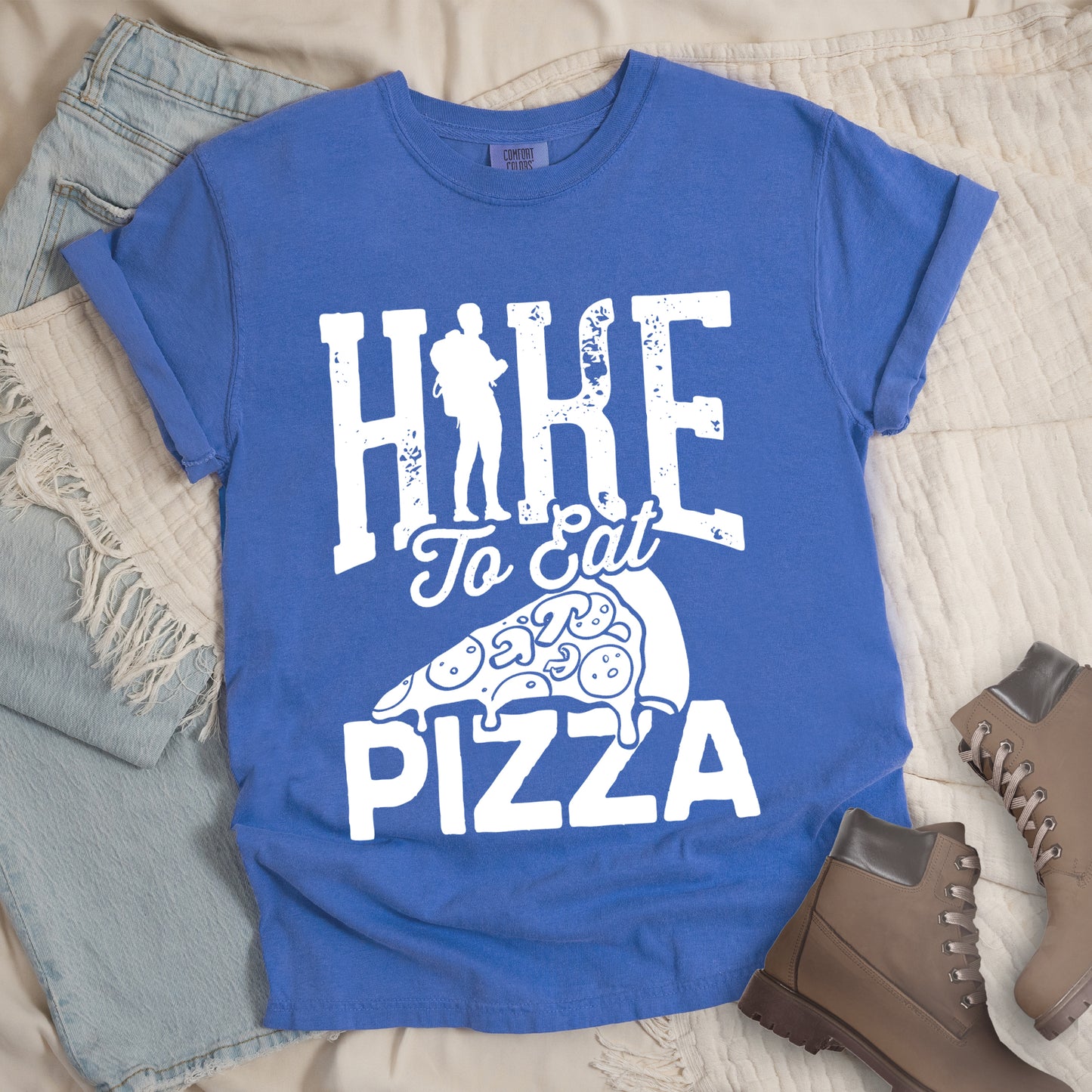 Flo Blue (Bright Blue) t-shirt with white distressed text reading "HIKE To Eat PIZZA". "HIKE" appears in large block letters at the top with a silhouette of a hiker with backpack integrated into the "I", "To Eat" in script in the middle, and "PIZZA" in large block letters at the bottom. Features a simple line drawing of a pizza slice with toppings between the phrases. The design humorously connects hiking activity with pizza as a reward.