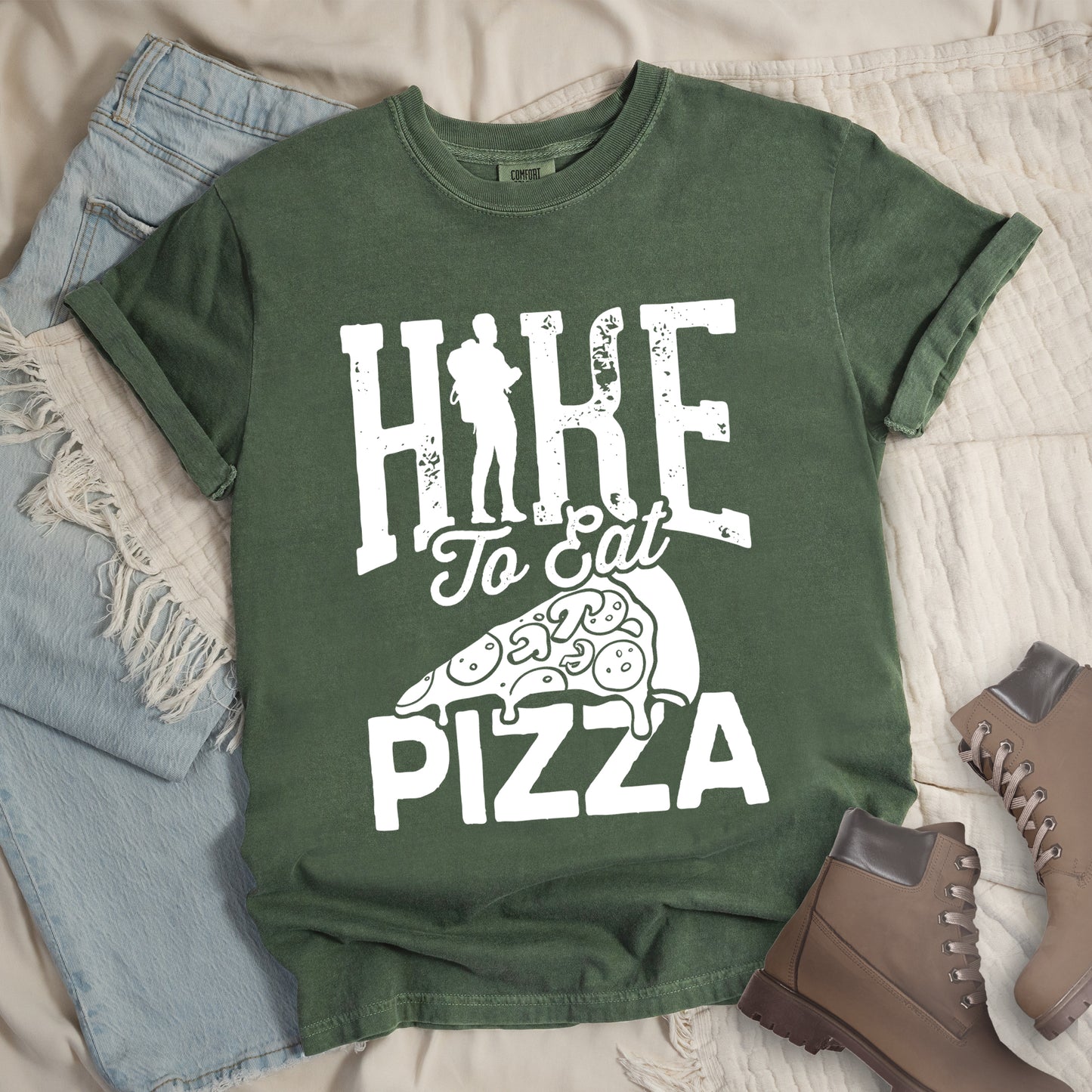 Moss (Dark Green) t-shirt with white distressed text reading "HIKE To Eat PIZZA". "HIKE" appears in large block letters at the top with a silhouette of a hiker with backpack integrated into the "I", "To Eat" in script in the middle, and "PIZZA" in large block letters at the bottom. Features a simple line drawing of a pizza slice with toppings between the phrases. The design humorously connects hiking activity with pizza as a reward.