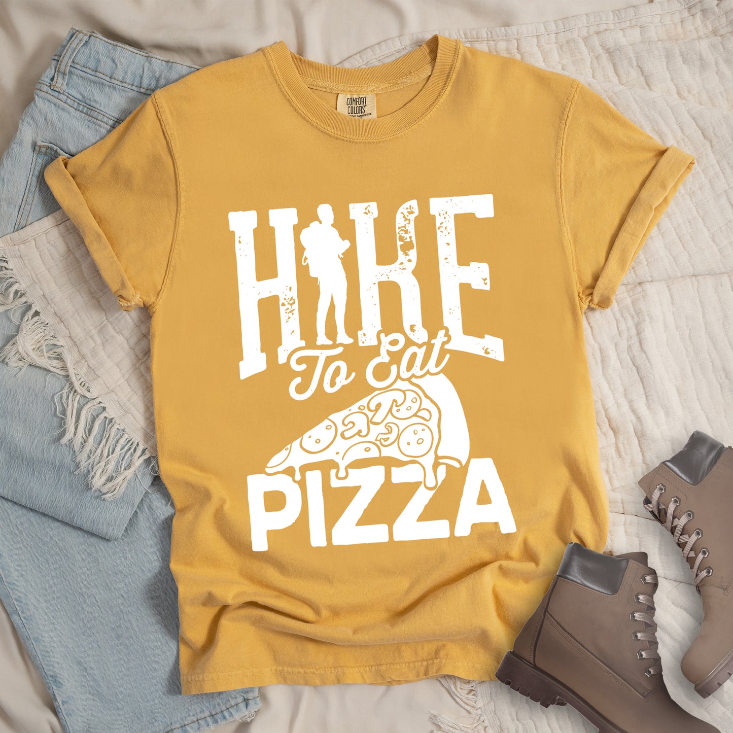 Mustard (Yellow) t-shirt with white distressed text reading "HIKE To Eat PIZZA". "HIKE" appears in large block letters at the top with a silhouette of a hiker with backpack integrated into the "I", "To Eat" in script in the middle, and "PIZZA" in large block letters at the bottom. Features a simple line drawing of a pizza slice with toppings between the phrases. The design humorously connects hiking activity with pizza as a reward.