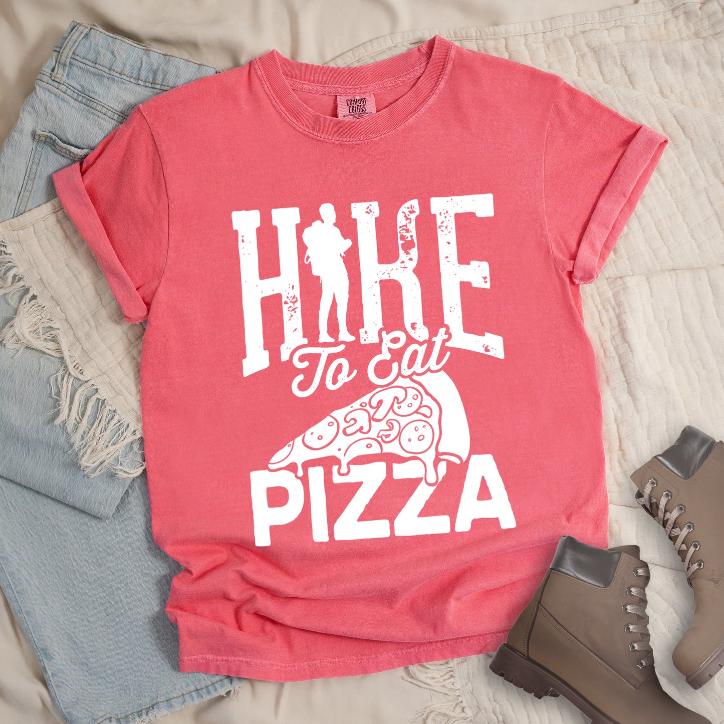 Watermelon (Light Coral) t-shirt with white distressed text reading "HIKE To Eat PIZZA". "HIKE" appears in large block letters at the top with a silhouette of a hiker with backpack integrated into the "I", "To Eat" in script in the middle, and "PIZZA" in large block letters at the bottom. Features a simple line drawing of a pizza slice with toppings between the phrases. The design humorously connects hiking activity with pizza as a reward.