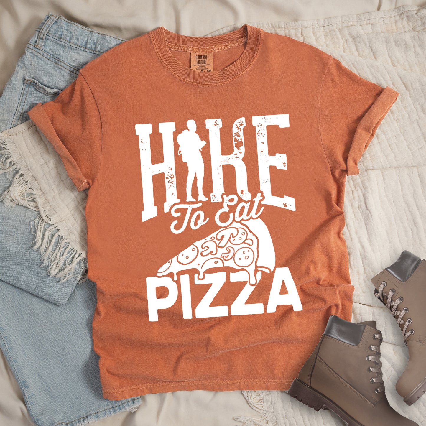 Yam (Brown) t-shirt with white distressed text reading "HIKE To Eat PIZZA". "HIKE" appears in large block letters at the top with a silhouette of a hiker with backpack integrated into the "I", "To Eat" in script in the middle, and "PIZZA" in large block letters at the bottom. Features a simple line drawing of a pizza slice with toppings between the phrases. The design humorously connects hiking activity with pizza as a reward.