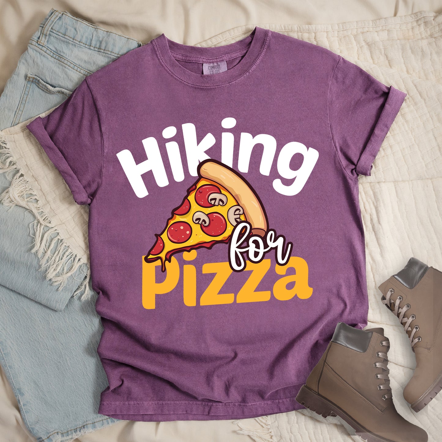 Berry (Dark Violet) t-shirt with white and orange text reading "Hiking for PIZZA". "Hiking" appears in white curved text at the top, "for" in small white script in the middle, and "PIZZA" in bold orange letters at the bottom. The centerpiece is a detailed illustration of a pizza slice with pepperoni and mushroom toppings on a yellow cheese background with tan crust. Features a classic crew neck and casual rolled sleeves, styled alongside light wash denim and brown hiking boots.