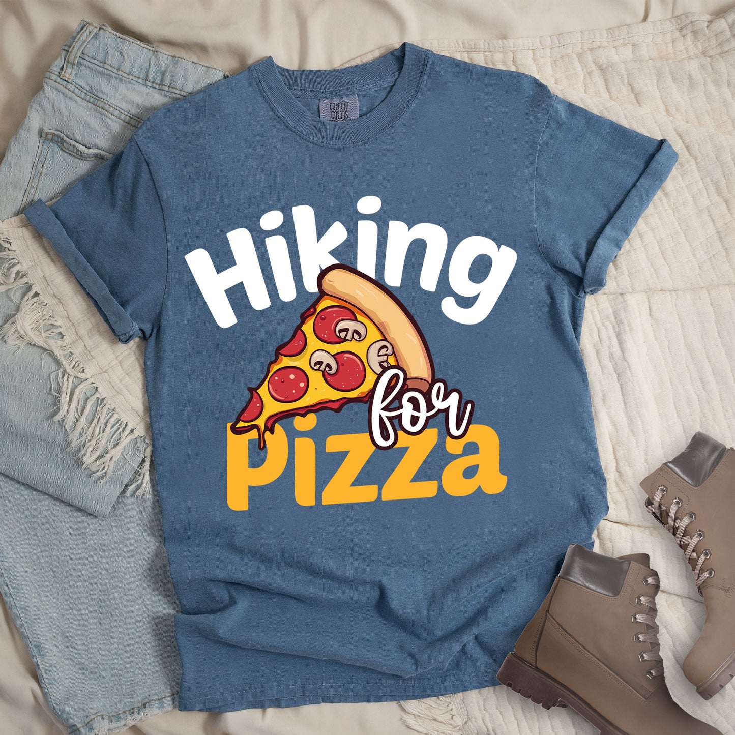 Blue Jean (Denim Blue) t-shirt with white and orange text reading "Hiking for PIZZA". "Hiking" appears in white curved text at the top, "for" in small white script in the middle, and "PIZZA" in bold orange letters at the bottom. The centerpiece is a detailed illustration of a pizza slice with pepperoni and mushroom toppings on a yellow cheese background with tan crust. Features a classic crew neck and casual rolled sleeves, styled alongside light wash denim and brown hiking boots.
